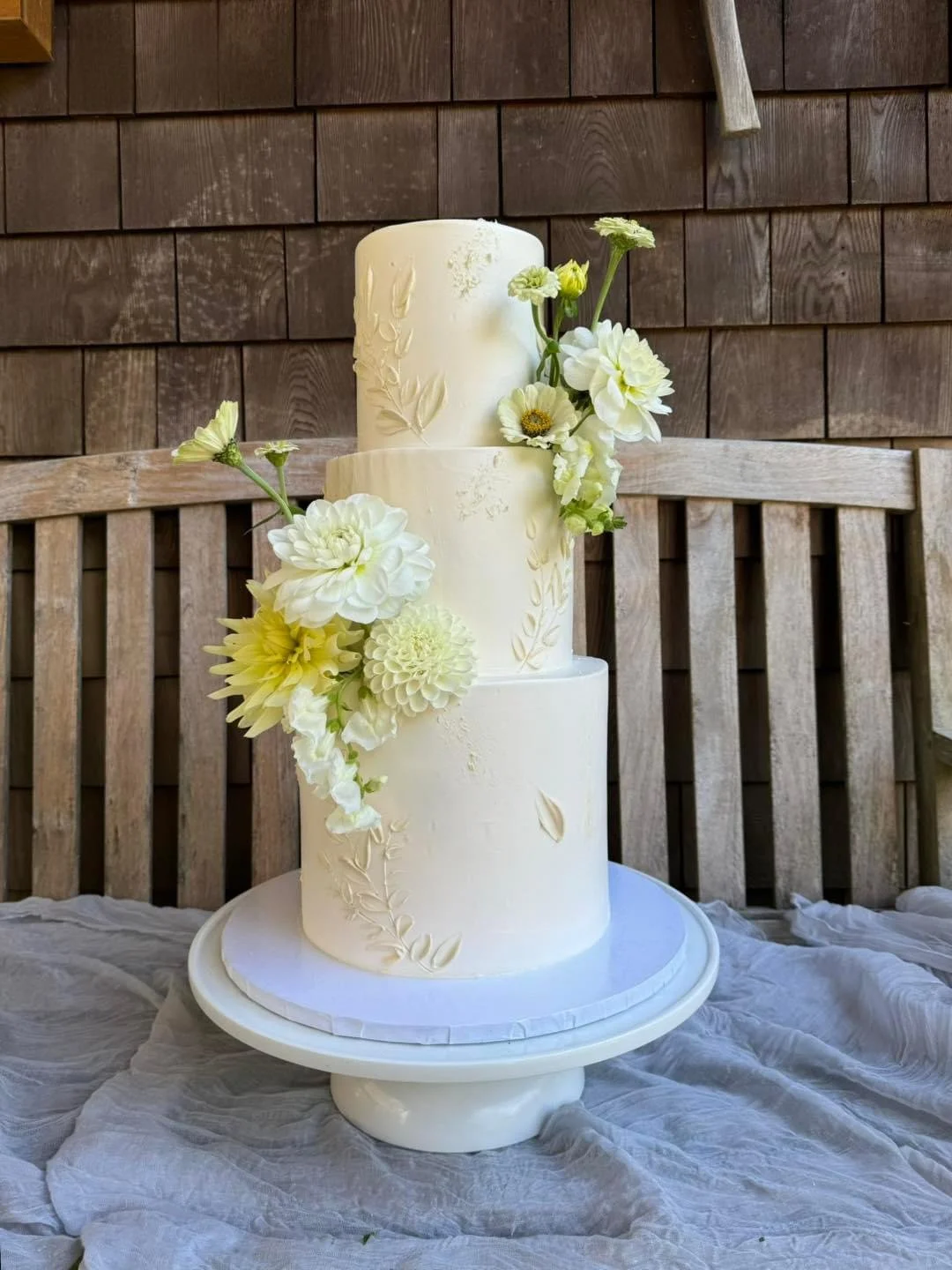 Three-tier white wedding cake decorated with white flowers and subtle embossed floral designs, placed on a white cake stand with a draped fabric underneath, against a wooden slat background.