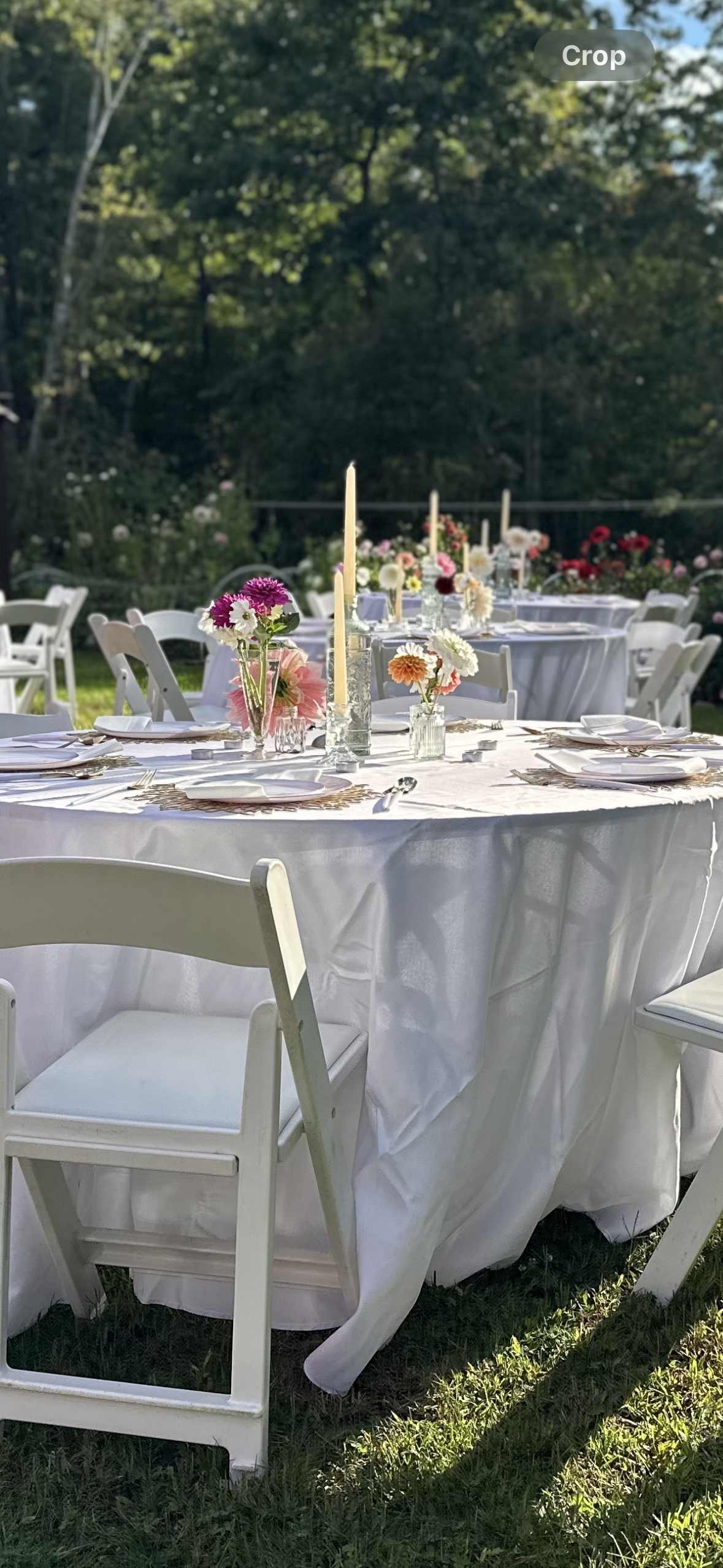 Outdoor wedding or event reception tables decorated with flowers and tall candles, set on a grassy area with trees in the background.