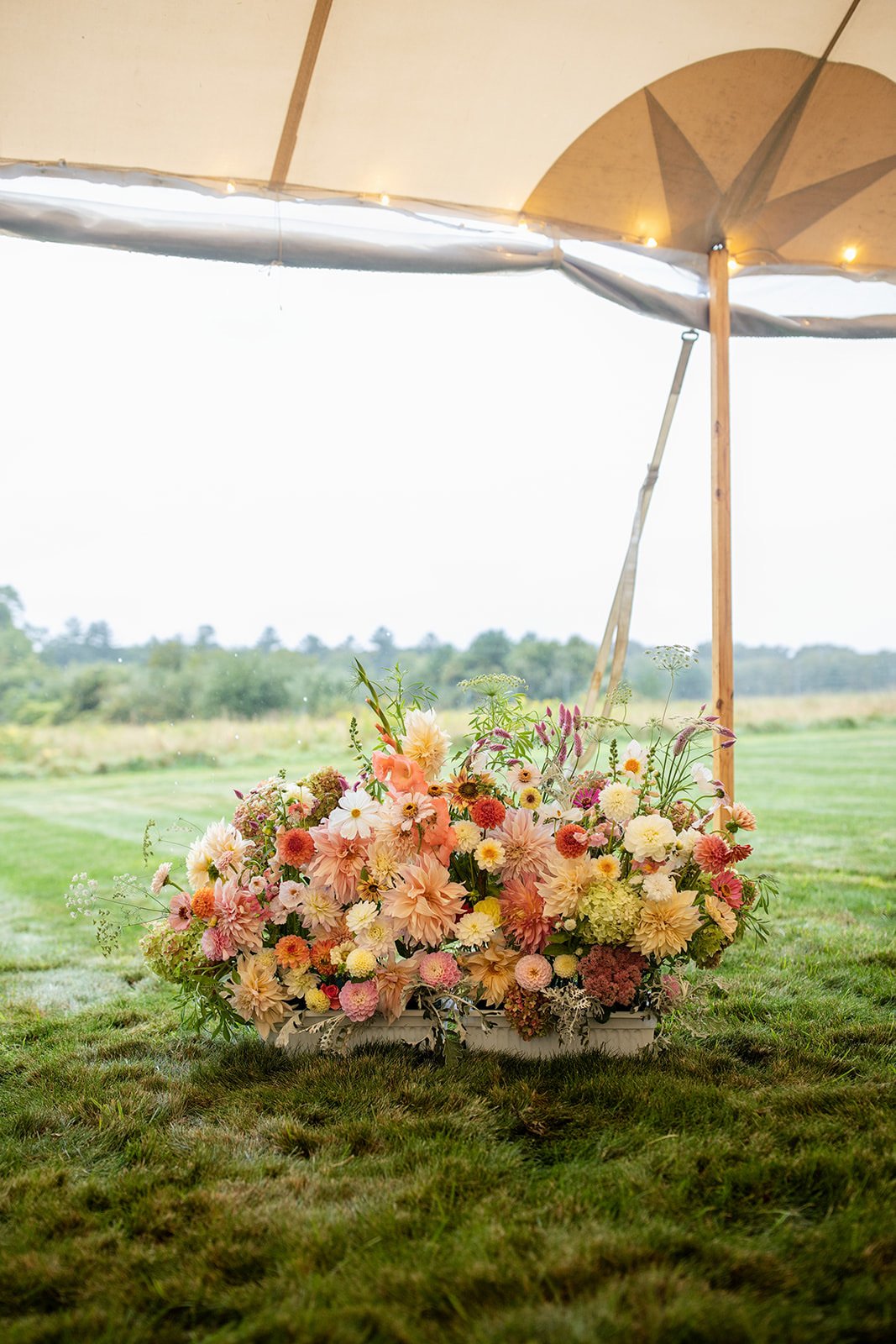 A large floral arrangement of pink, peach, cream, and white flowers under a wooden and beige umbrella in an outdoor grassy field.