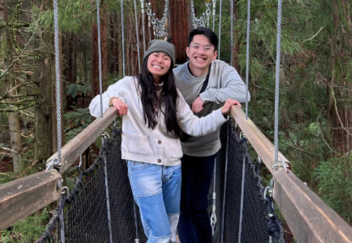 Two people, a woman and a man, smiling on a suspension bridge in a forest, with tall trees and greenery around.