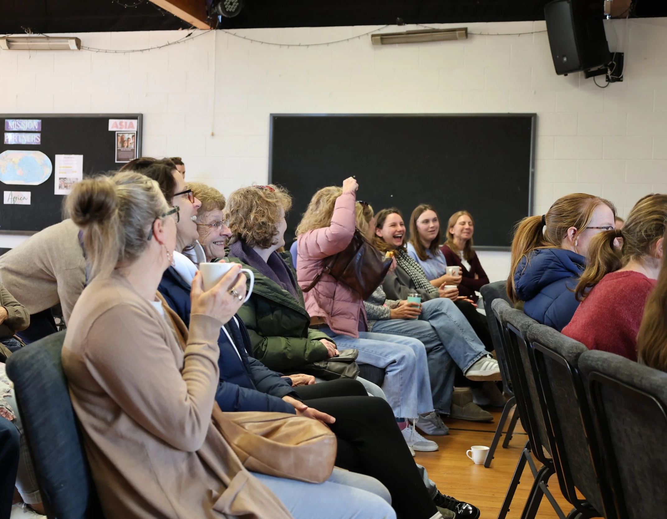 A diverse group of people sitting in a classroom, laughing and enjoying a presentation or lecture. They are seated in rows with notebooks and cups, and the classroom features blackboards, speakers, and a world map on the wall.