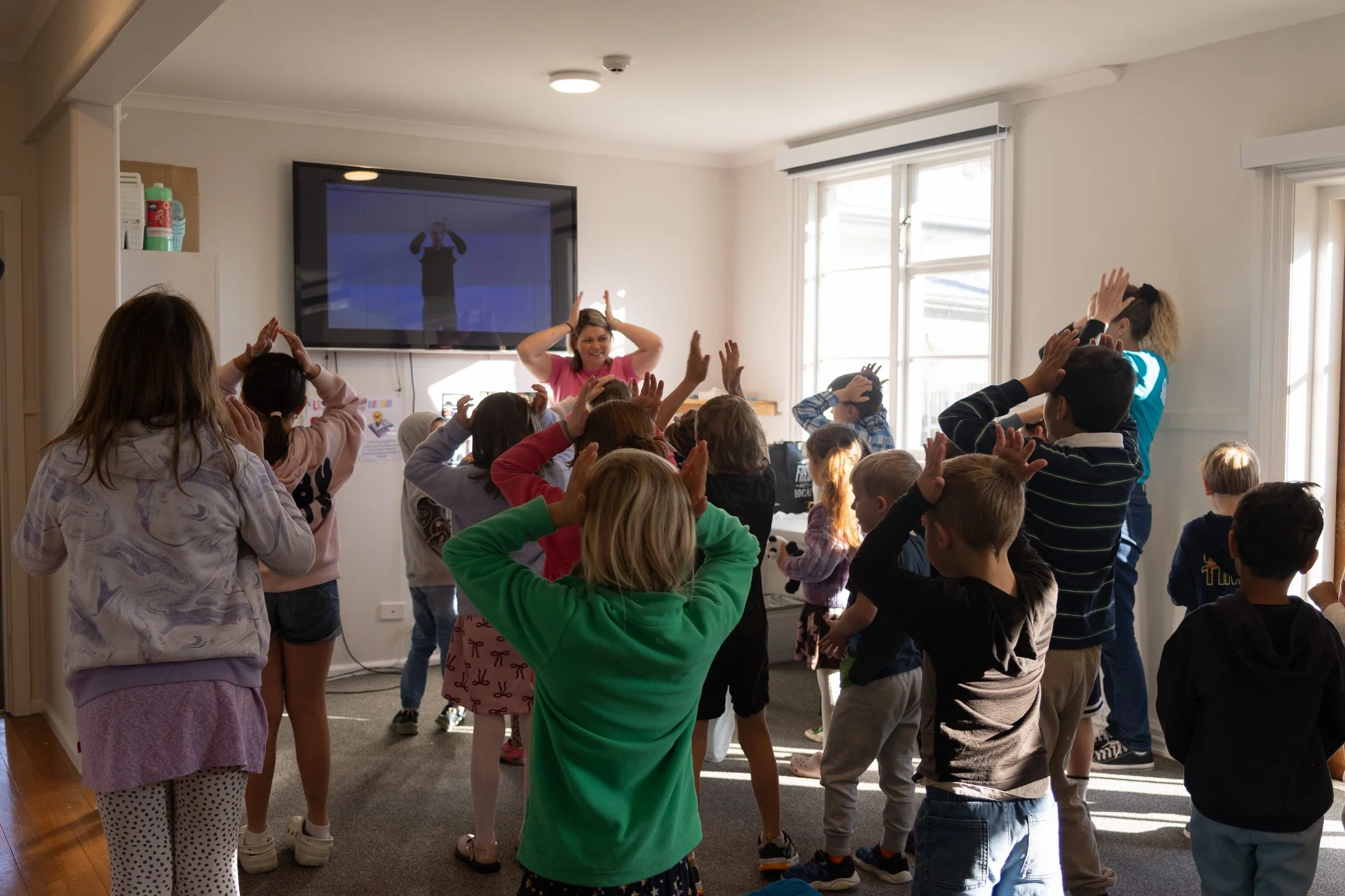 Children standing in a classroom, raising their hands and participating in an activity, with a woman leading them.