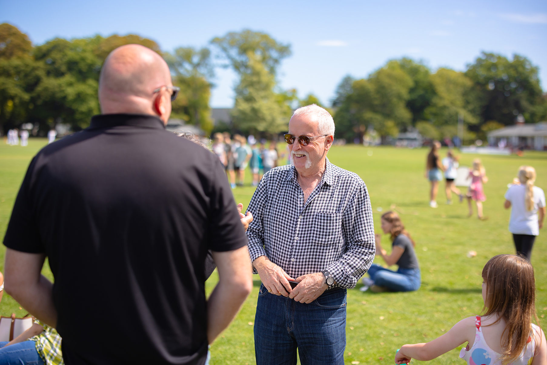 Two men with sunglasses shaking hands and talking at an outdoor event on a sunny day, with children and adults in the background on a green field with trees.