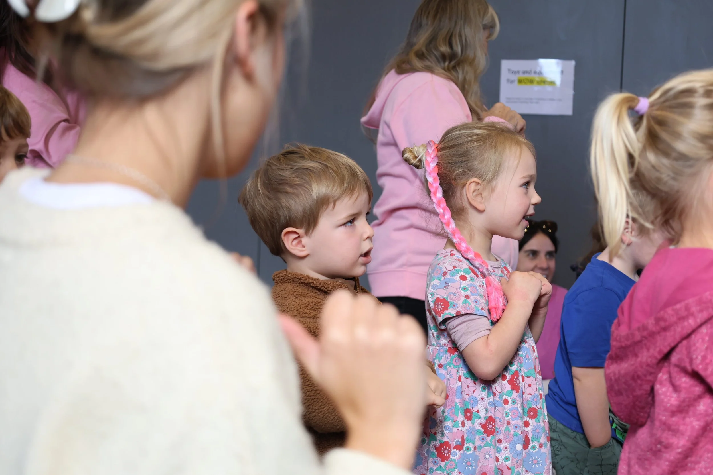 Children and adults standing in a line indoors, possibly singing or participating in an activity, with some clapping and engaged expressions.