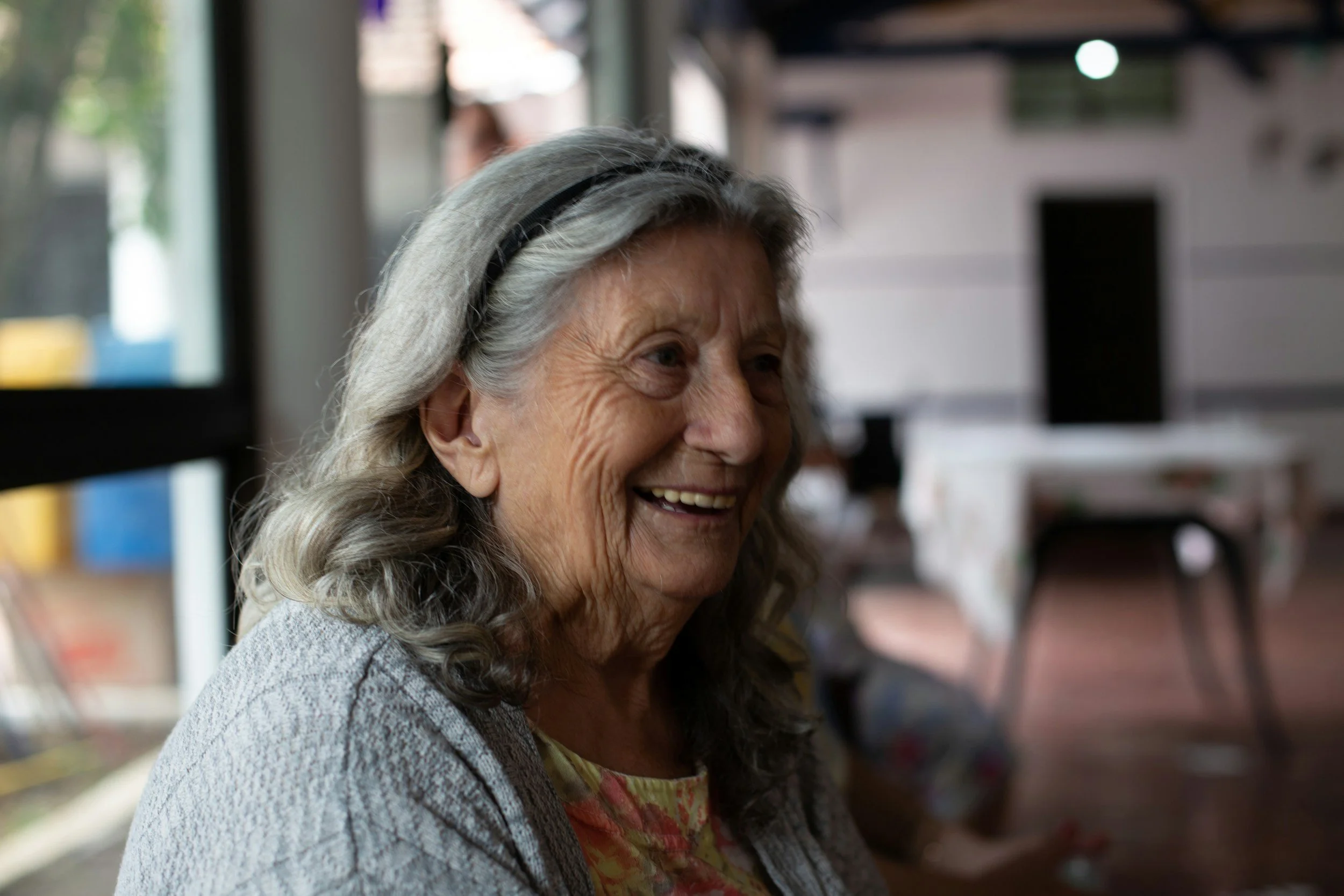 Smiling elderly woman with gray hair and a black headband, wearing a gray cardigan and floral shirt, indoors near a window.