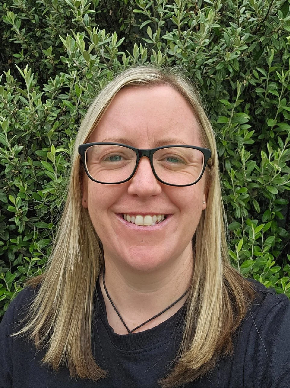 A woman with blonde hair, glasses, and a necklace smiling outdoors in front of green foliage.