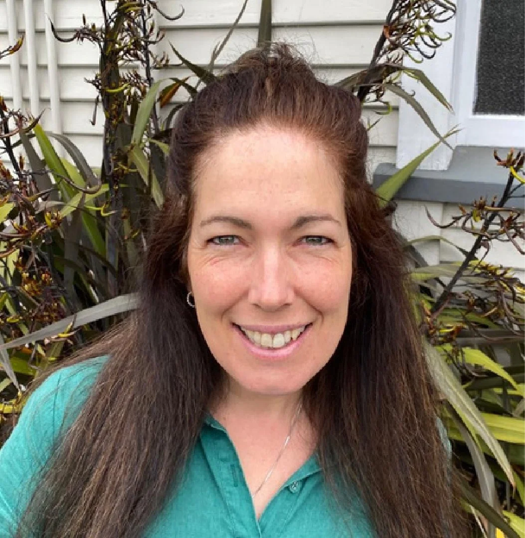 A woman with long brown hair smiling outdoors in front of plants and a house.