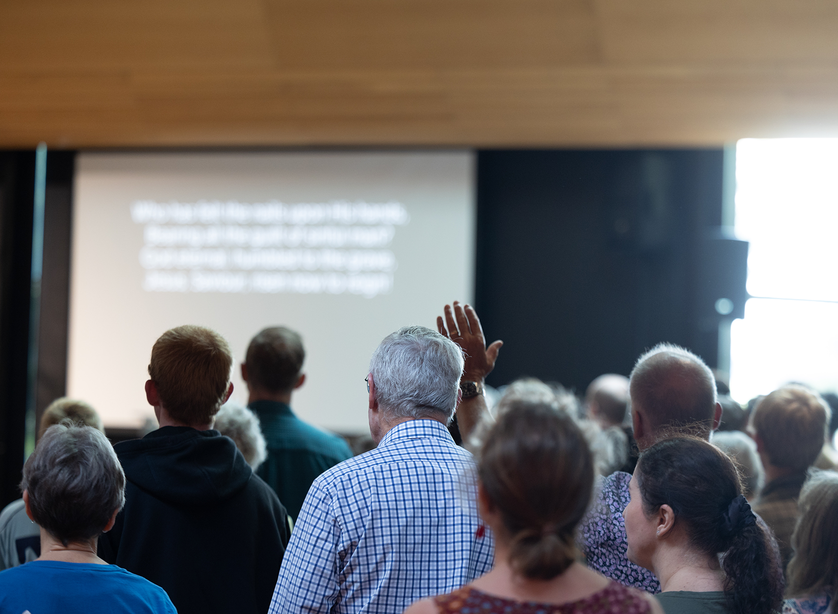 A crowd of people attending a presentation or lecture in a room with a large screen in the background. One person is raising their hand.