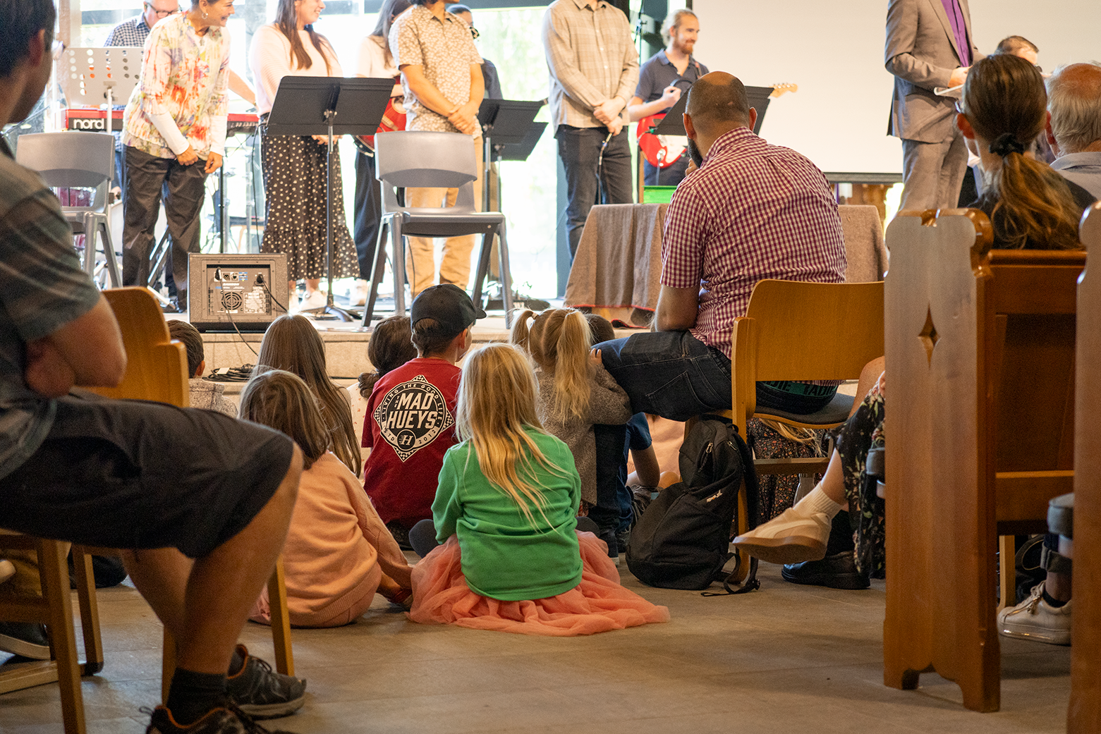 Children and adults sitting and watching a live musical performance in a community room, with the band performing on a stage at the front.
