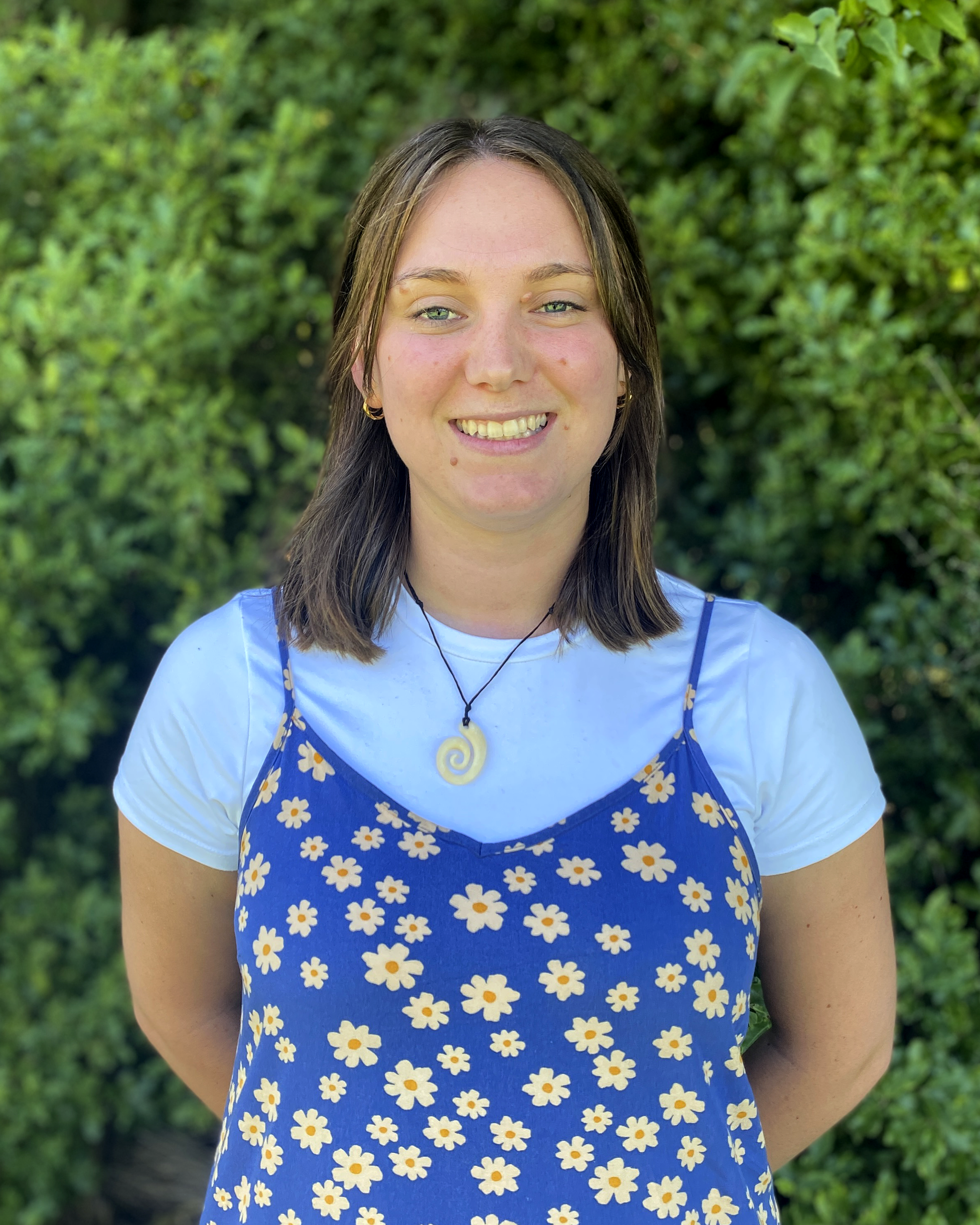 A young woman with light skin, shoulder-length brown hair, and green eyes, smiling, wearing a blue dress with white daisy flower pattern over a white t-shirt, standing outdoors with green foliage in the background.