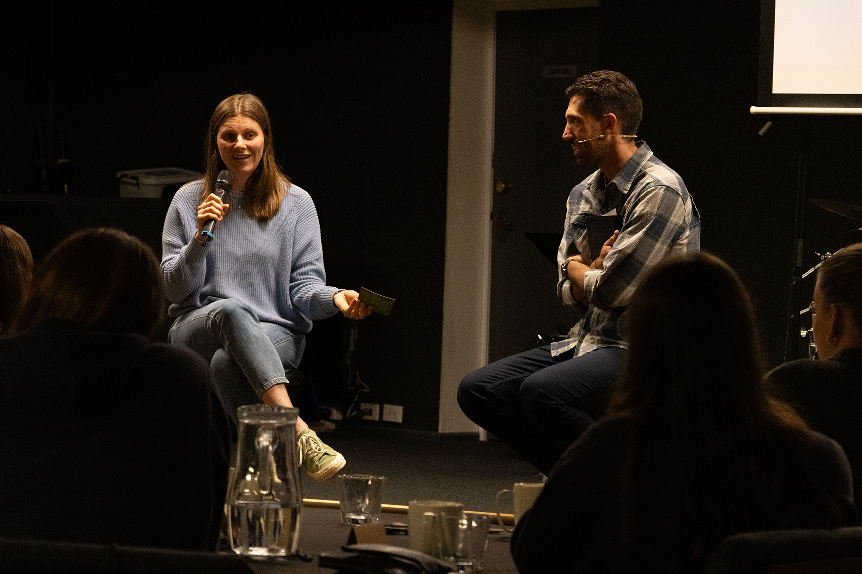 A young woman in a light blue sweater holding a microphone and a small note, sitting on a stool and speaking to an audience during a panel discussion. A man with dark hair, wearing a plaid shirt, is seated next to her, smiling and listening. The setting appears to be indoors, in a conference or event space, with several people in the audience.