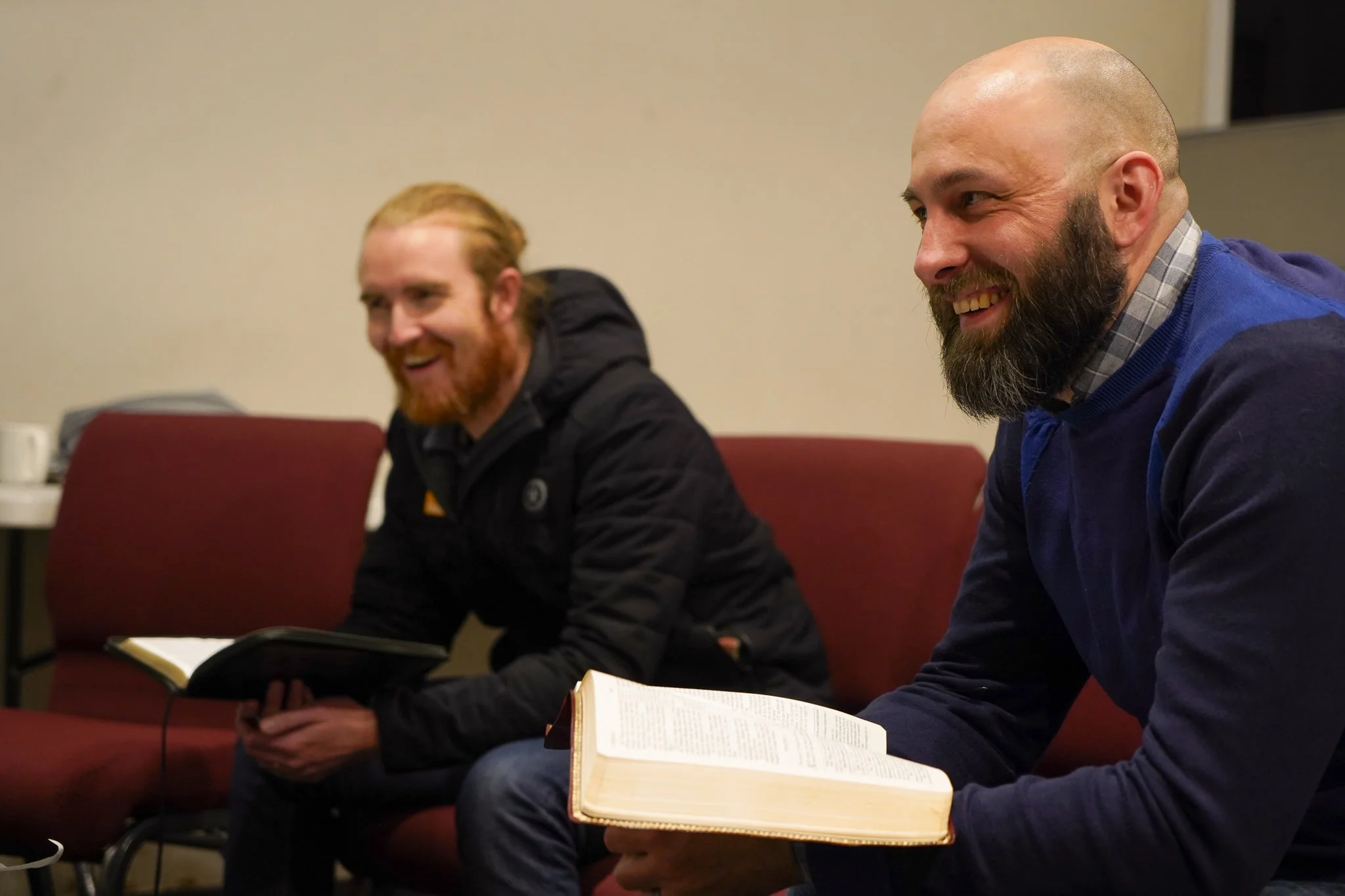Two men sitting on red chairs laughing during a meeting or discussion, with books and notebooks in front of them.