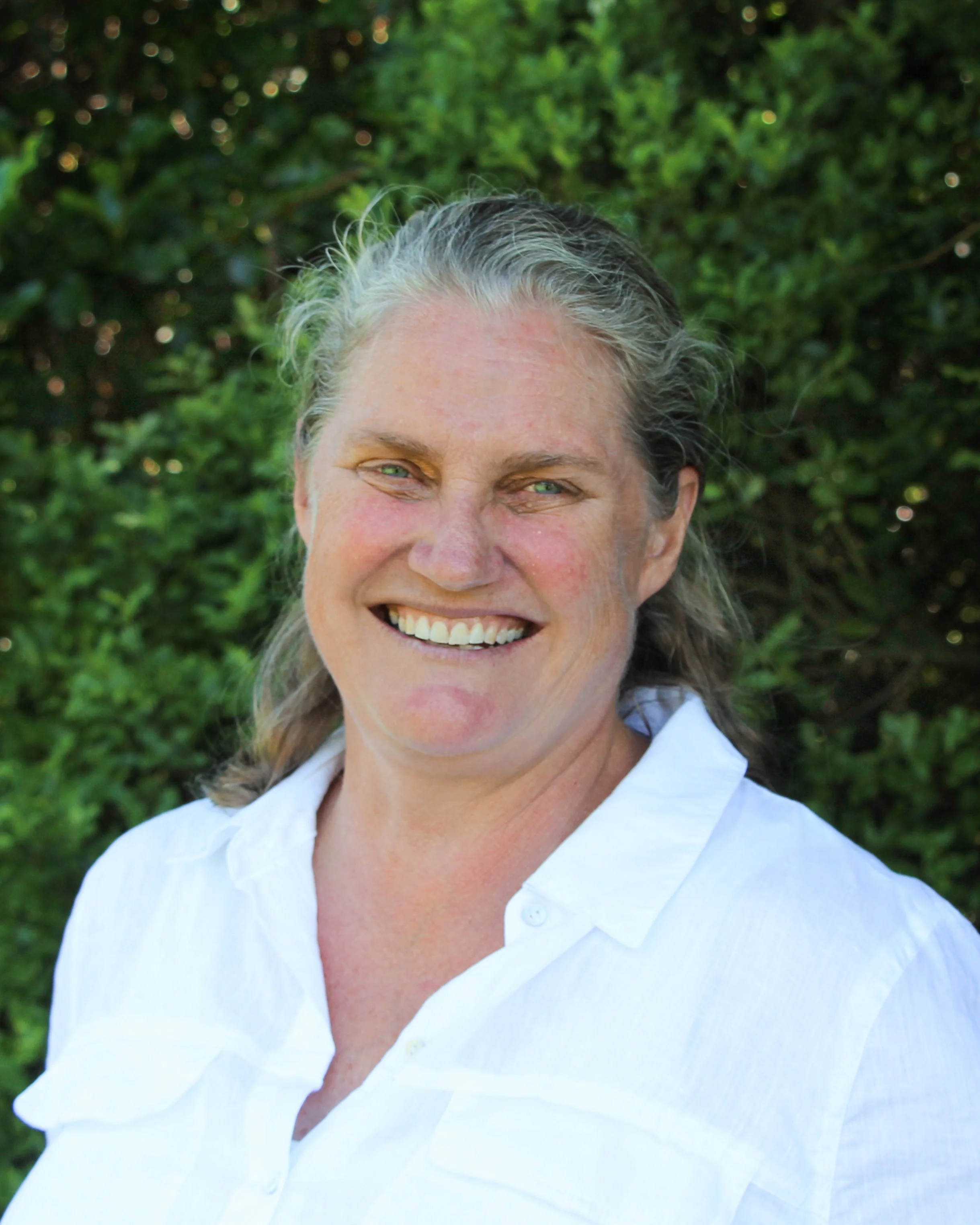 Close-up of a smiling woman with gray hair wearing a white shirt, outdoors with green foliage in the background.