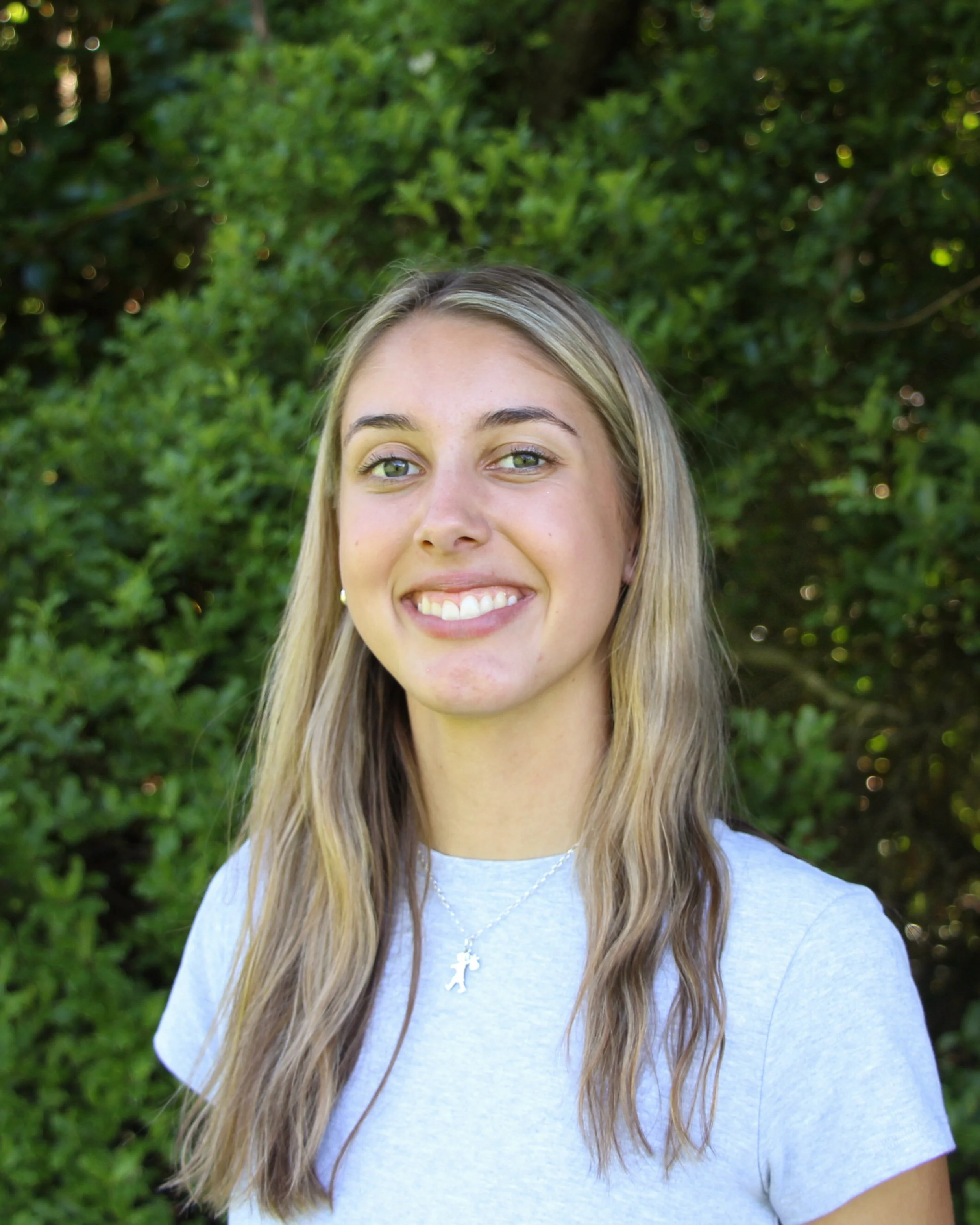 A young woman with long blonde hair smiling outdoors with green bushes in the background, wearing a light gray t-shirt and a cross necklace.