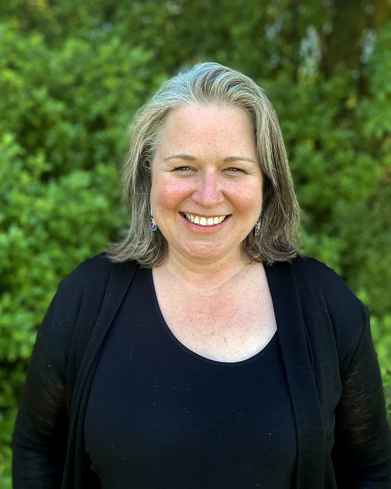 A woman smiling outdoors with green foliage in the background, wearing a black top and earrings.