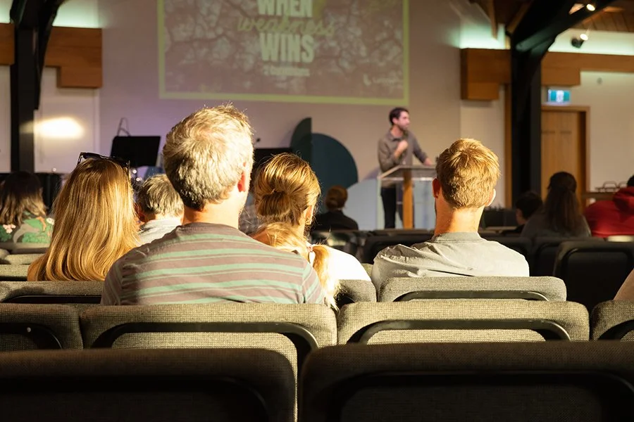 Audience seated in a conference room watching a speaker on stage, with a presentation slide projected behind him.