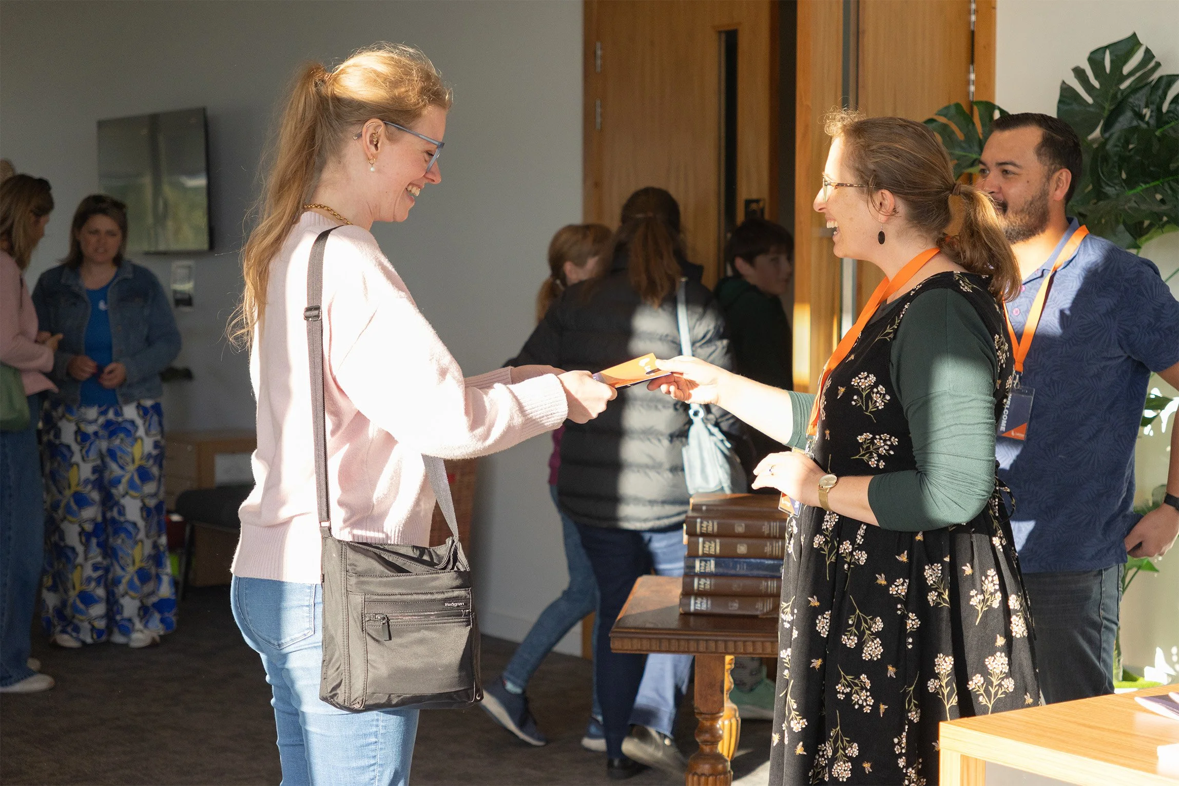 A woman with glasses and long hair, wearing a pink sweater, receives an award or certificate from another woman with glasses, wearing a black floral dress, who is smiling. Several people are in the background at an indoor event.