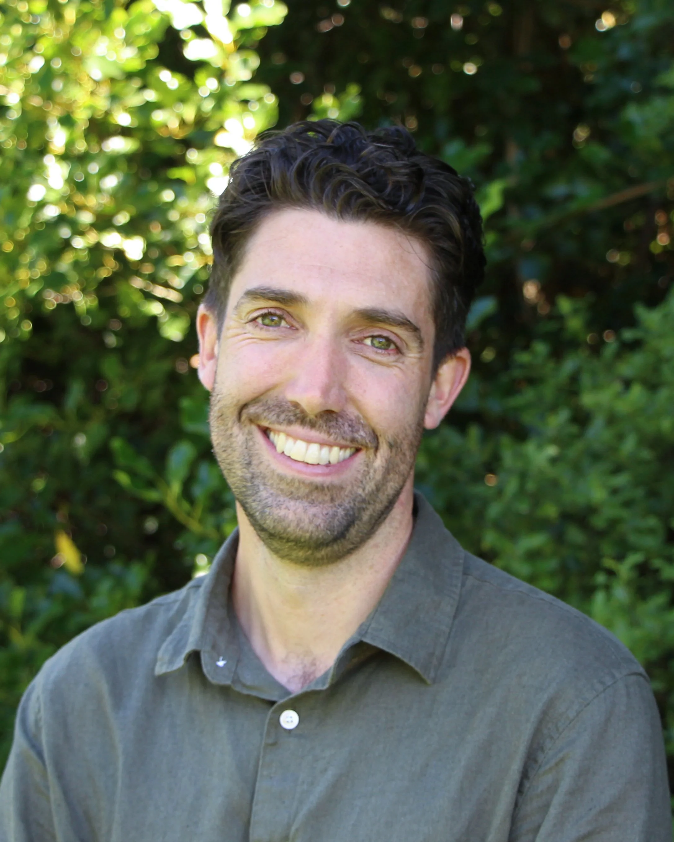 Close-up of a smiling man with dark wavy hair, light skin, wearing a gray button-up shirt, standing outdoors with green foliage in the background.