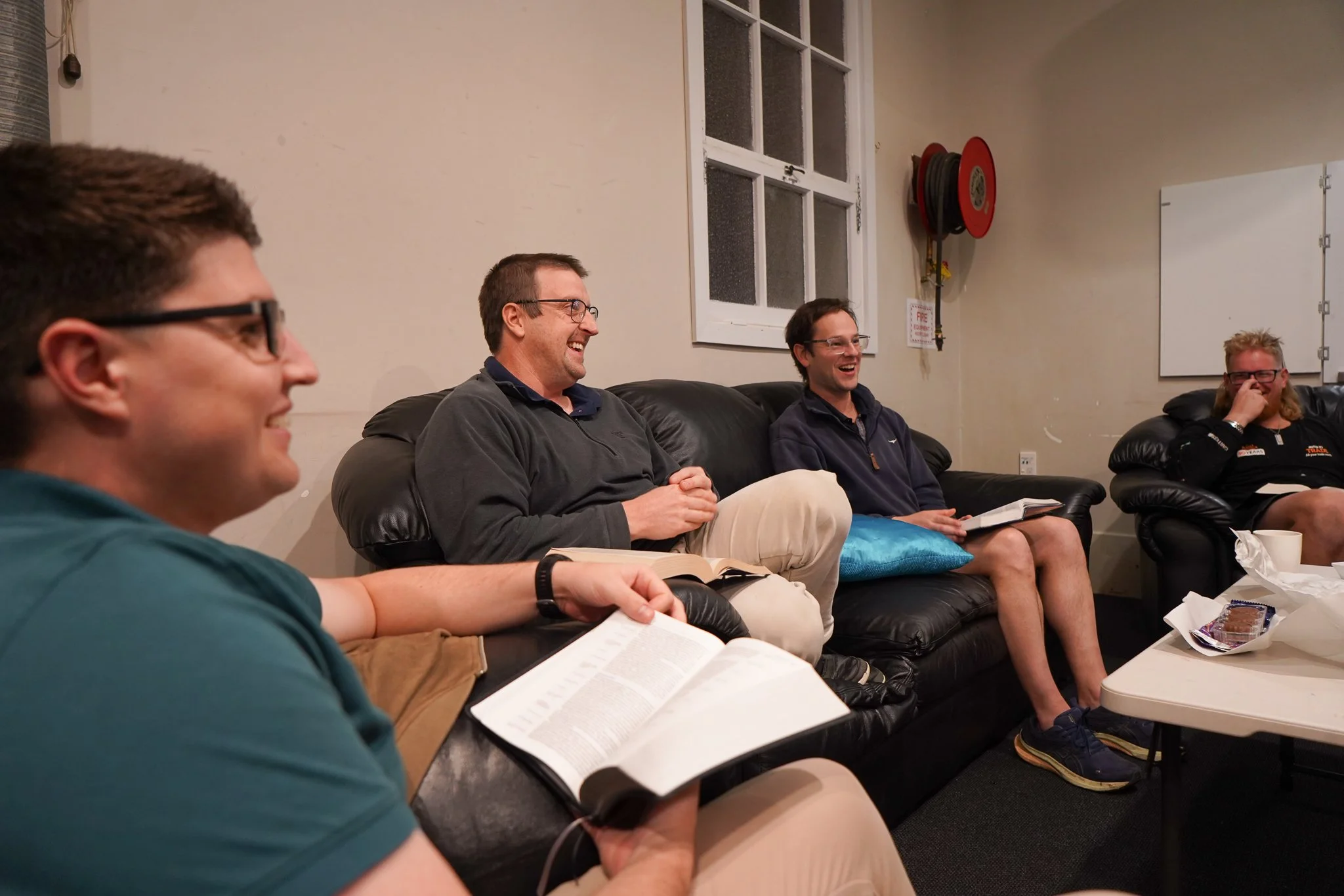Four people sitting on a black leather couch, laughing and talking in a room with white walls and large windows, with snacks and drinks on a table in front.