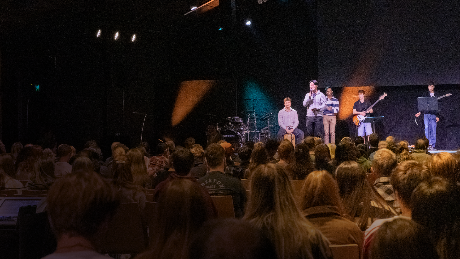 Group of people on stage performing and speaking in front of a large audience in a dark theater with stage lighting.