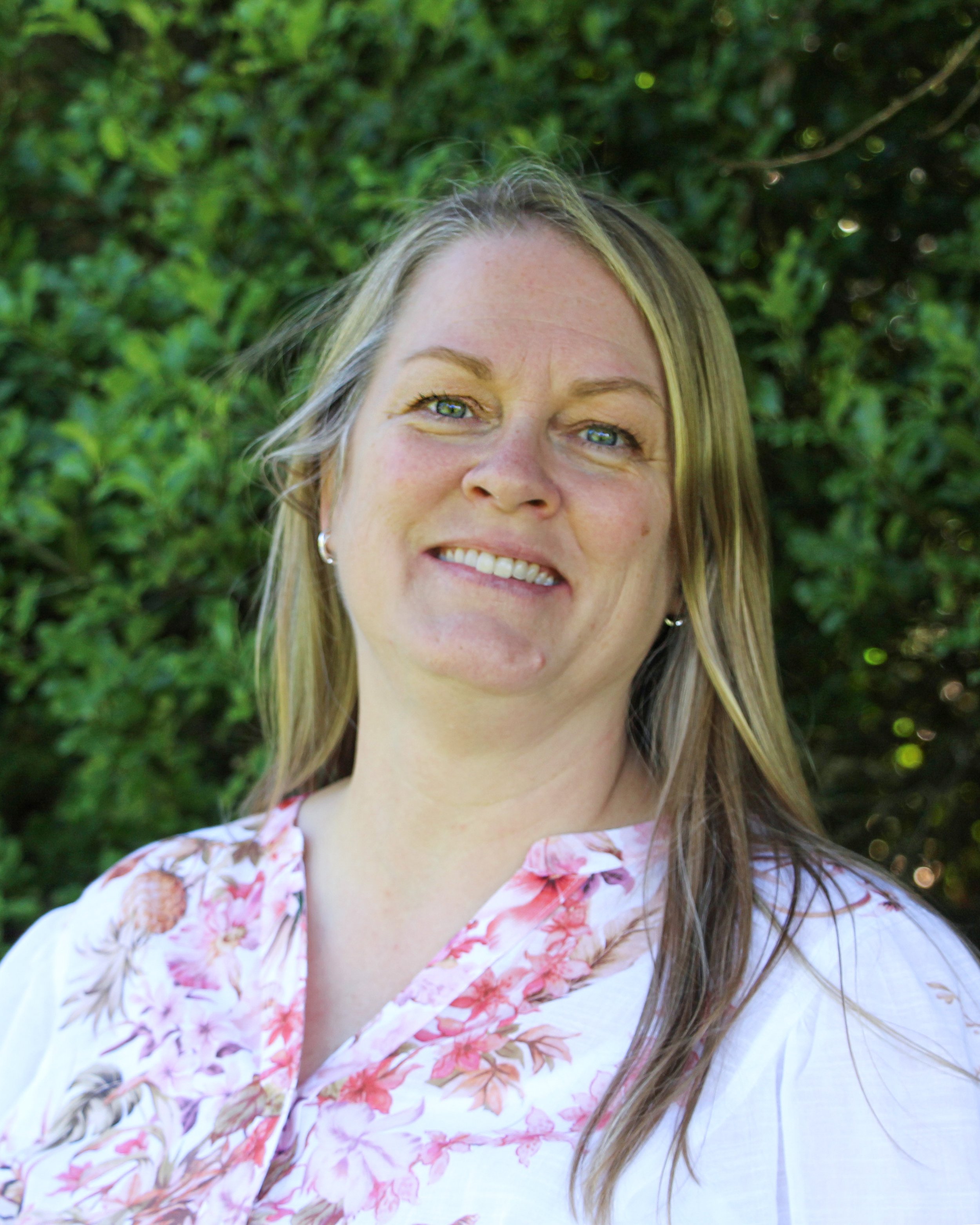 Close-up of a woman with blonde hair and blue eyes smiling, wearing earrings and a floral shirt, outdoors with greenery in the background.