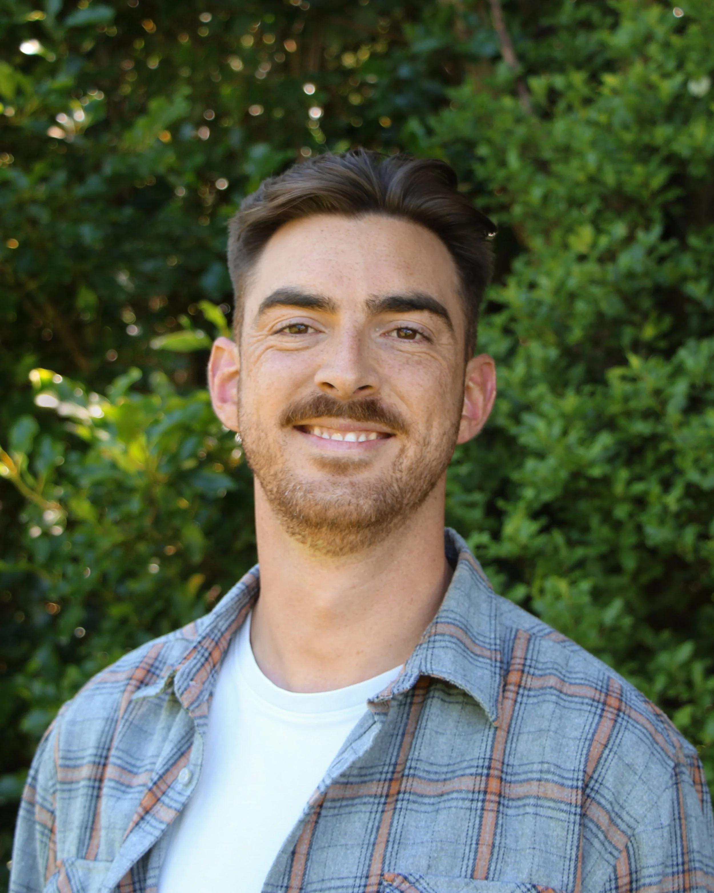 A smiling man with brown hair and a beard, wearing a white t-shirt and a blue plaid shirt, standing outdoors with green foliage in the background.