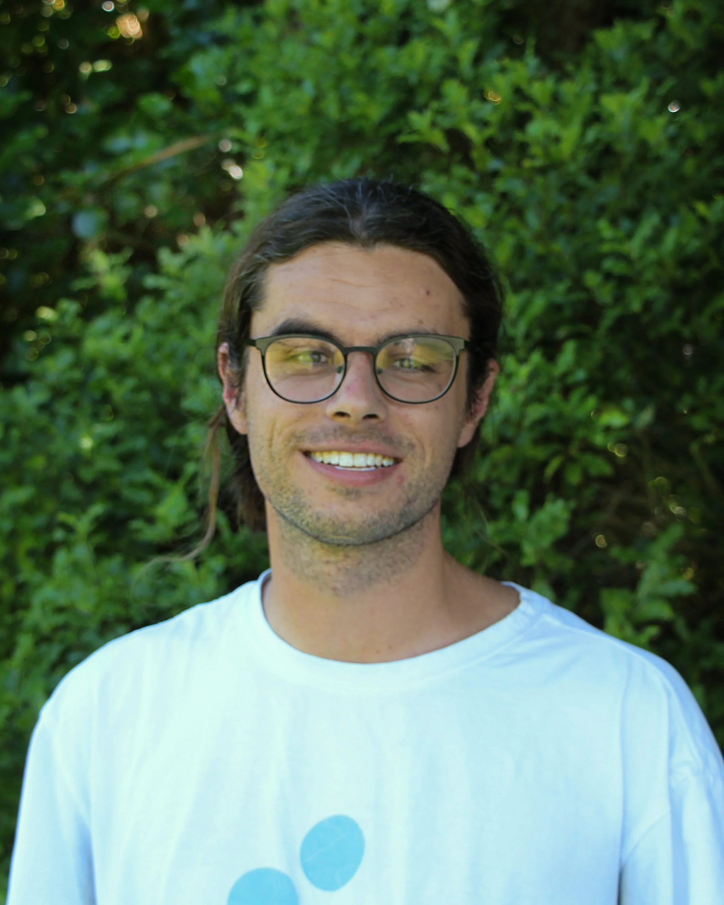 A young man with long brown hair, glasses, and light skin, smiling outdoors in front of green foliage, wearing a white T-shirt with blue circles.