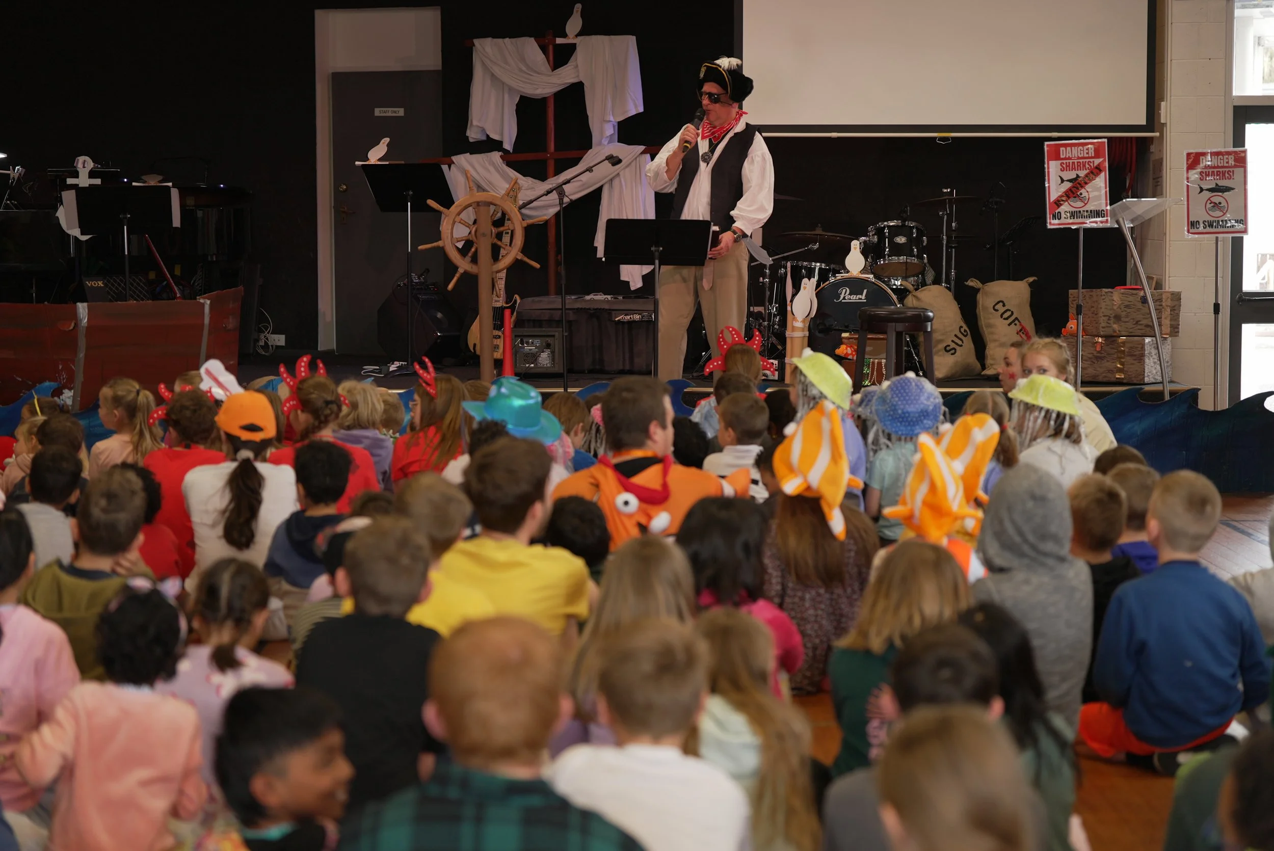 A man dressed as a pirate, speaking into a microphone on stage with children sitting in front, many of whom are wearing costumes like reindeer antlers and colorful hats.