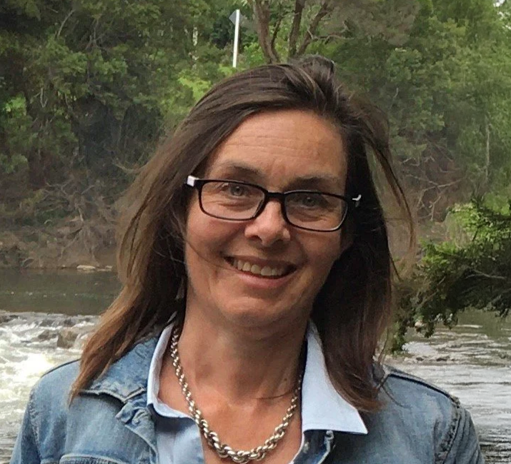 A woman smiling outdoors near a river with green trees in the background.