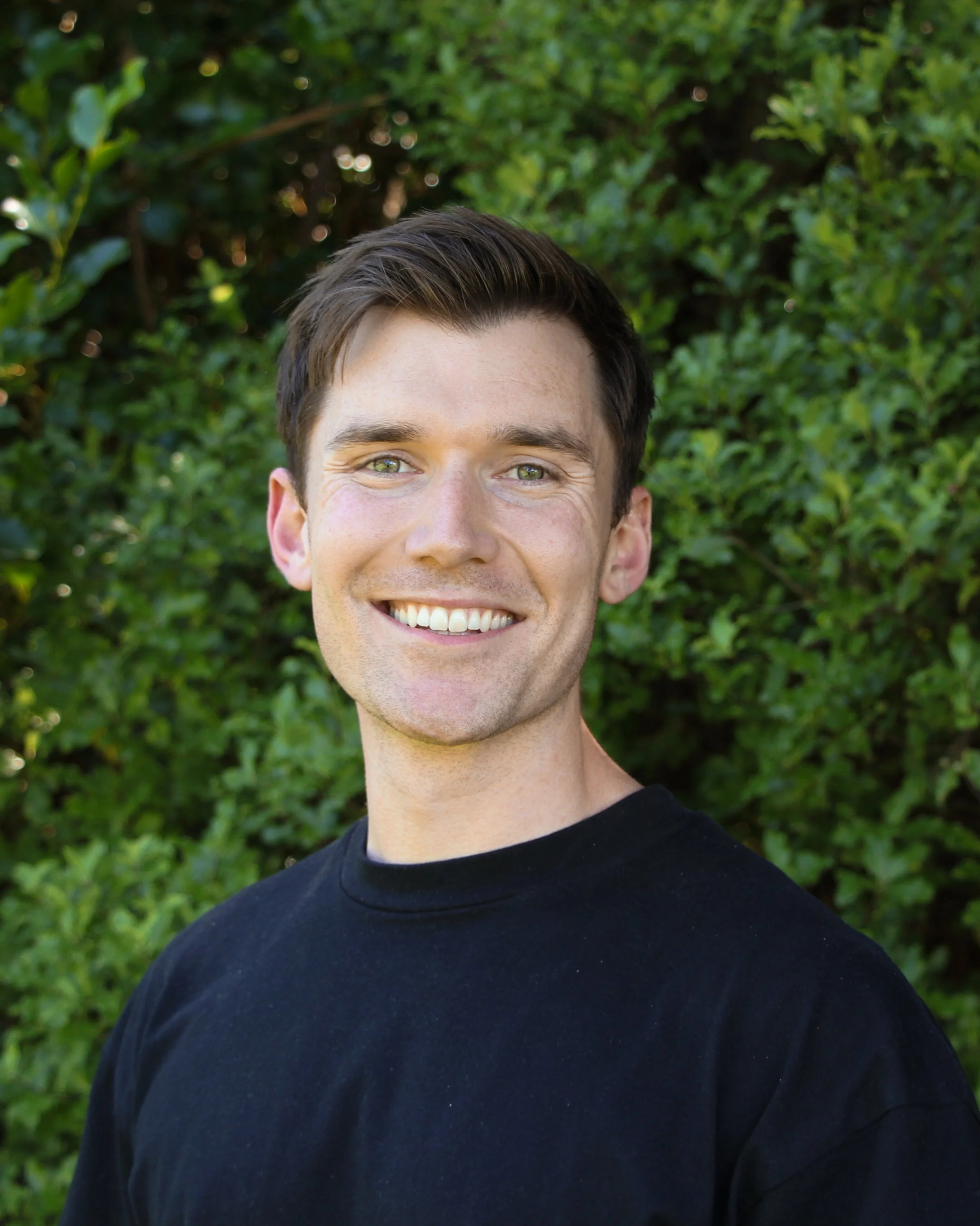 Close-up of a young man with short dark hair and green eyes, smiling and wearing a black shirt, standing outdoors in front of green foliage.