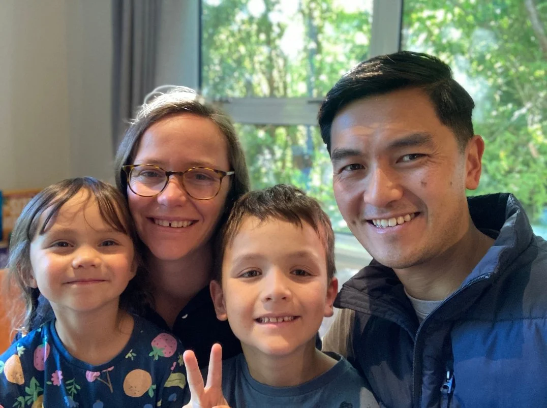 A family of four taking a selfie indoors with a window showing green trees outside, smiling and dressed casually.