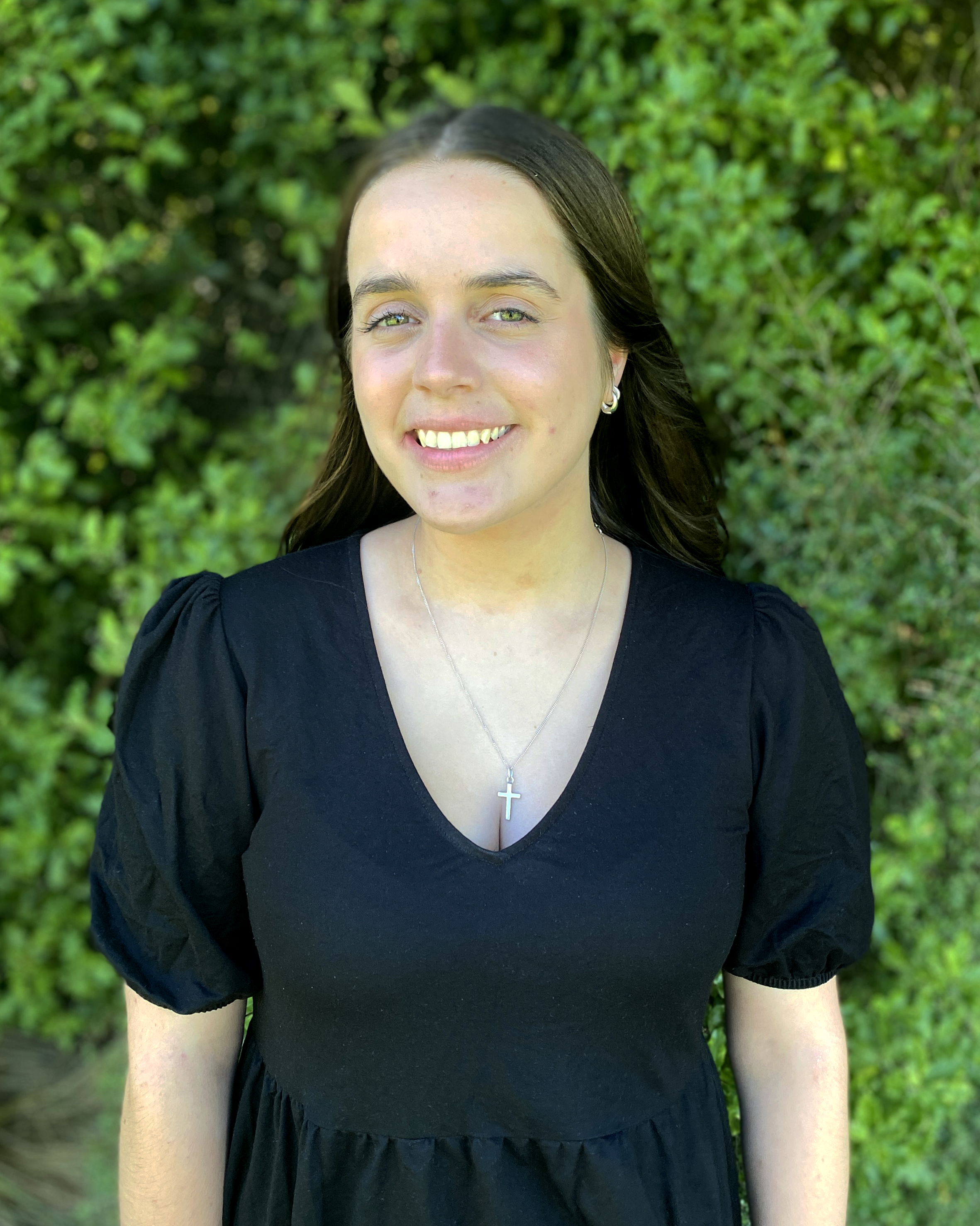 Close-up of a young woman with brown hair, wearing a black top, earrings, and a cross necklace, standing outdoors in front of green foliage, smiling at the camera.