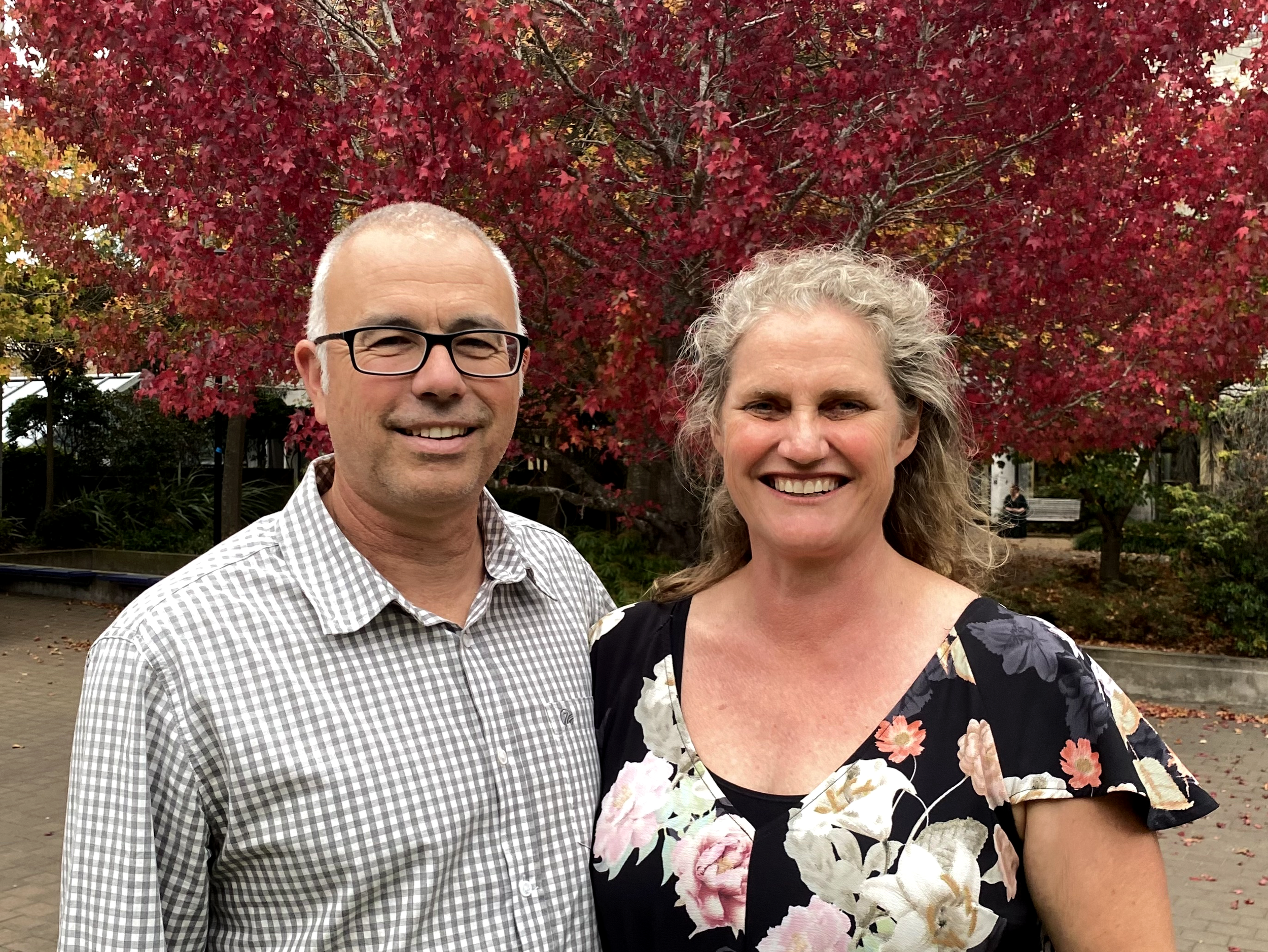 A smiling man wearing glasses and a checkered shirt standing next to a smiling woman with curly hair and a floral dress, outdoors in front of a red-leafed tree.