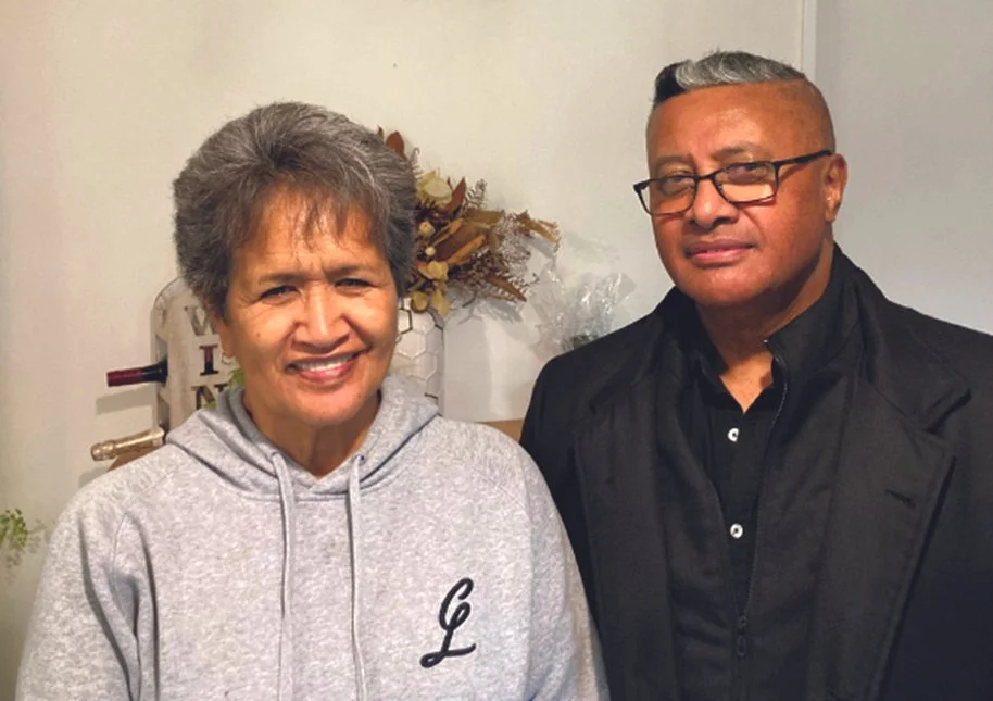 A smiling older woman with short gray hair wearing a light gray hoodie, standing next to a man with glasses and short gray hair, dressed in a black jacket and black shirt, in an indoor setting with neutral-colored walls and some decorative items in the background.