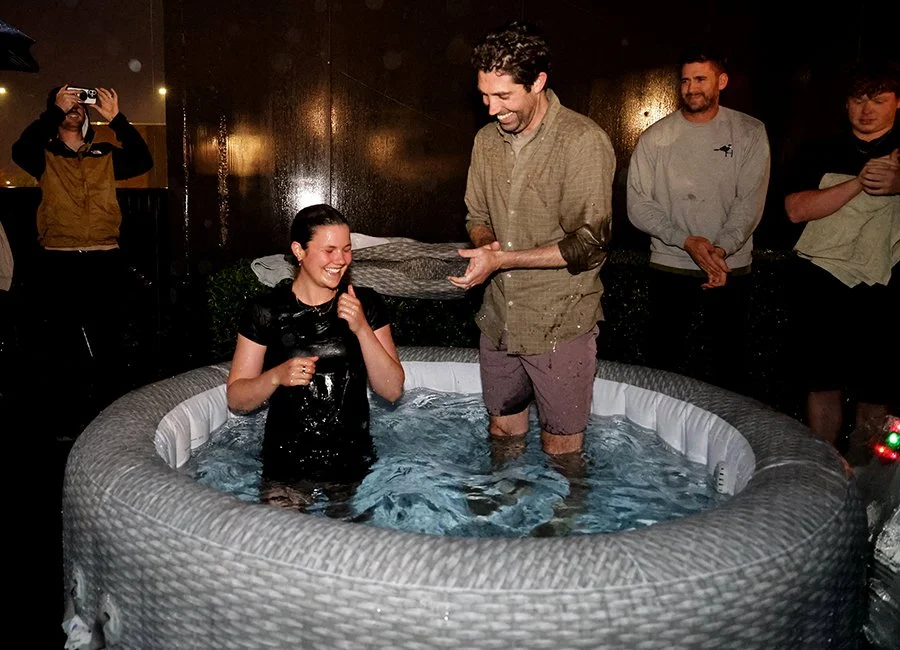 Woman getting baptized in a small indoor pool with a man holding a gray towel, surrounded by onlookers smiling and taking photos.