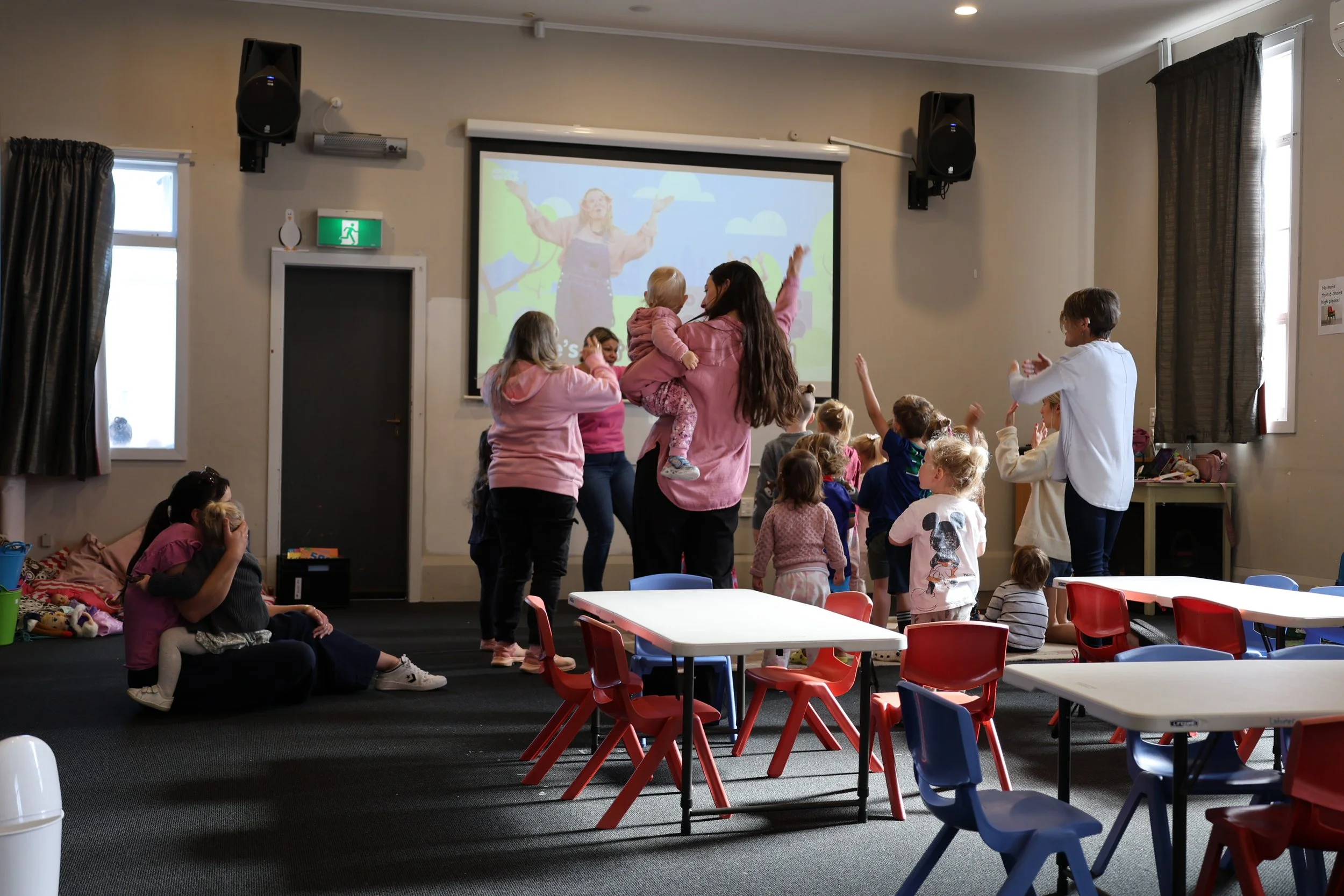 Children and adults in a classroom watching a presentation on a large screen, with some children sitting and some standing, raising their hands or interacting.