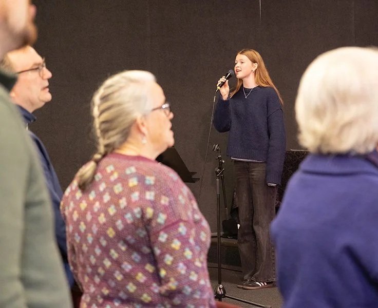A young woman with red hair singing into a microphone on a stage, with an audience in the foreground.
