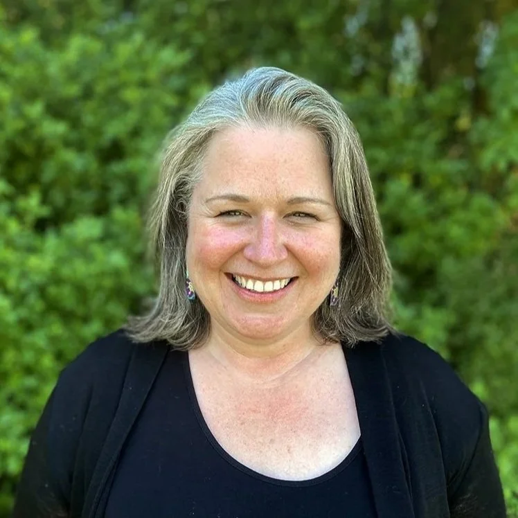 A smiling woman with shoulder-length gray hair, wearing earrings and a black top, standing outdoors with green foliage in the background.