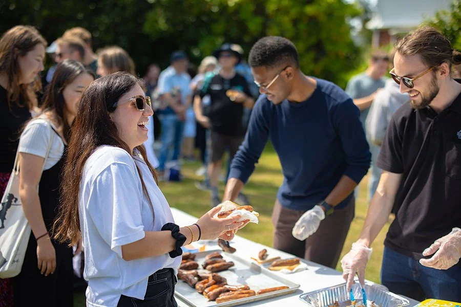 People gathered outdoors at a food service table, with a woman smiling and holding a hot dog, as others serve food behind the table.