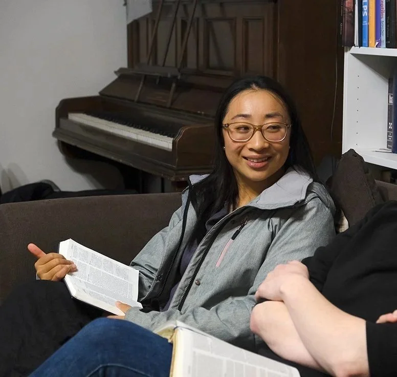 A woman with glasses sitting on a sofa, holding an open book, smiling and talking to someone.