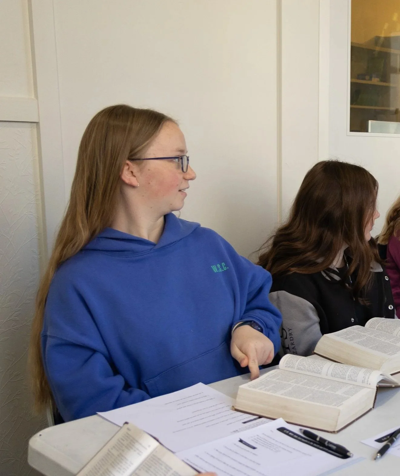 A young woman with long red hair, glasses, and a blue hoodie sitting at a table with open books, papers, and a pen, in a room with white walls.