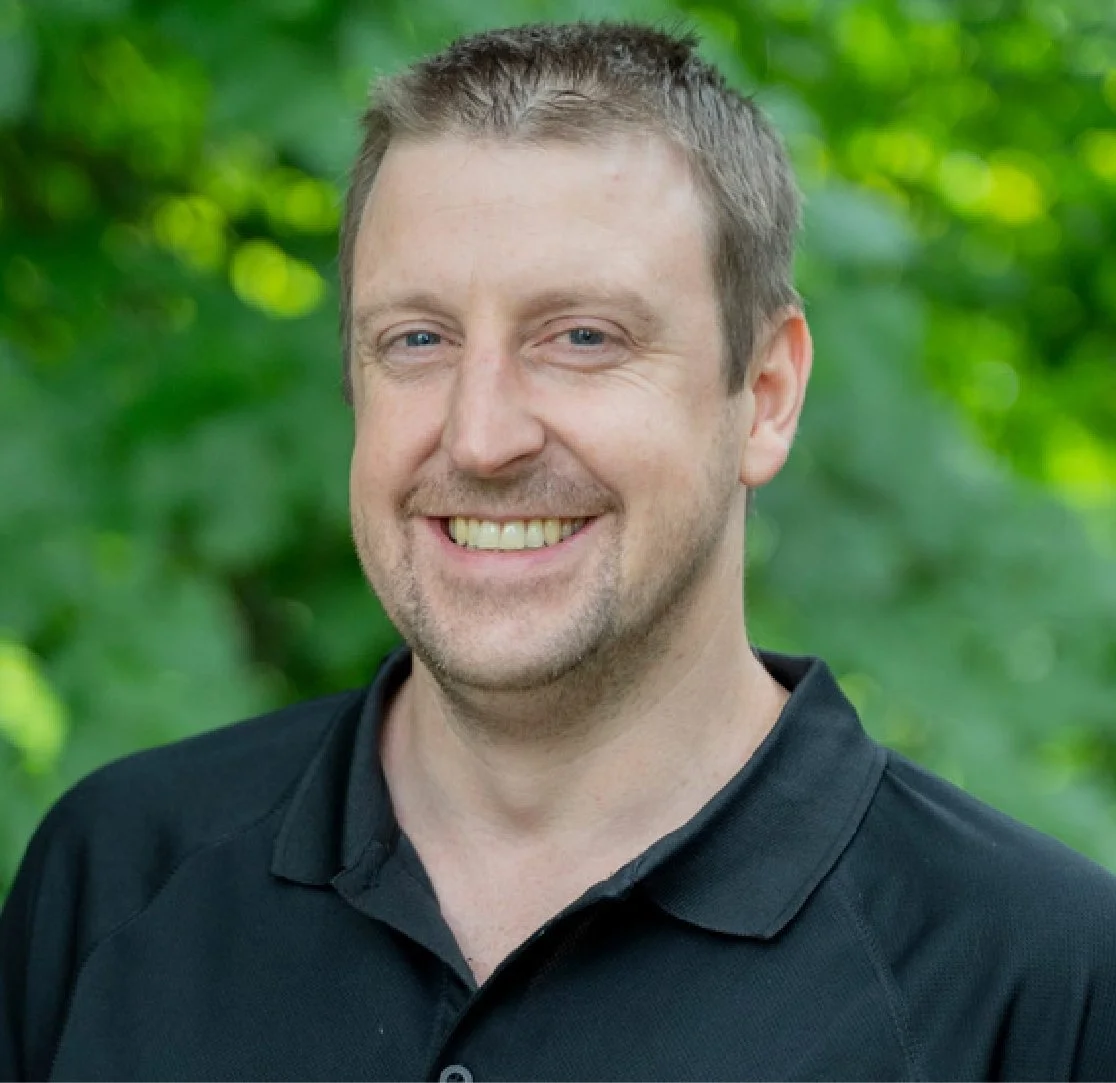 A smiling man with short brown hair and blue eyes, wearing a black polo shirt, standing outdoors with green foliage in the background.