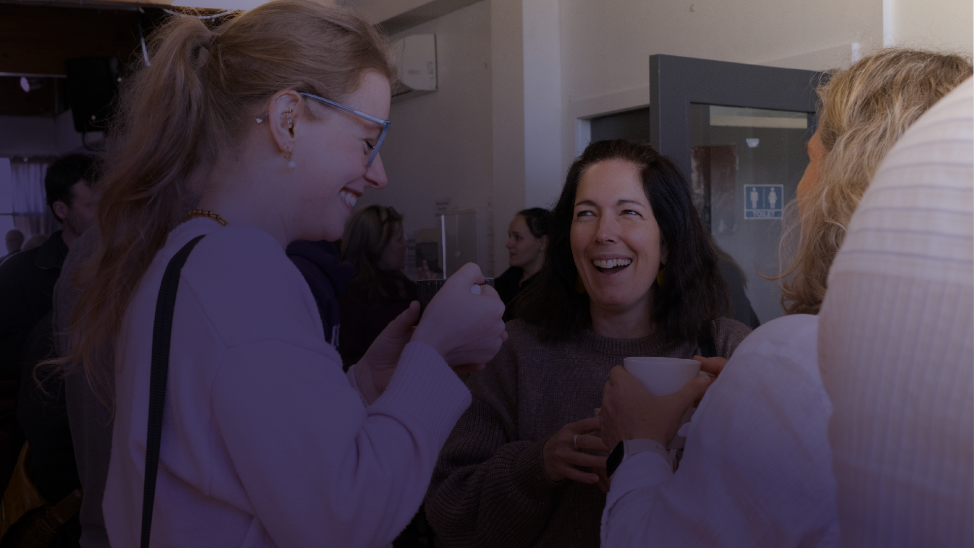 Group of women at a social gathering, smiling, holding drinks and engaging in conversation inside a building.