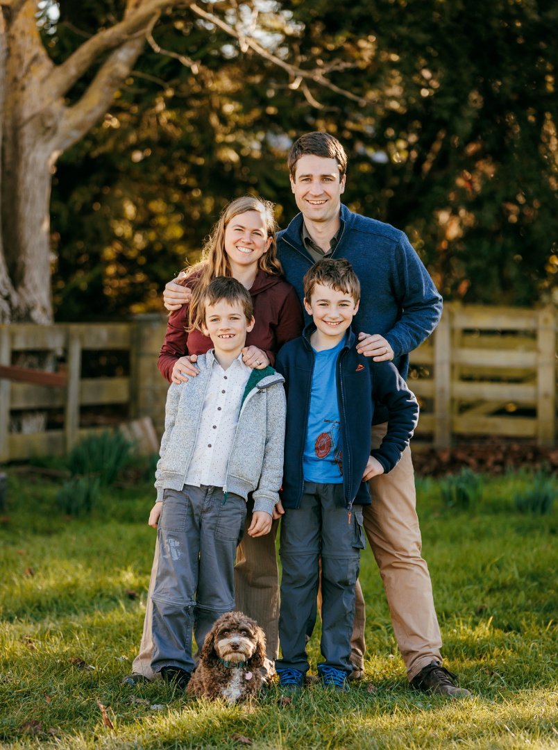 A family of four with two boys and two adults standing on grass outdoors, smiling and posing for the camera, with a dog sitting in front of them.