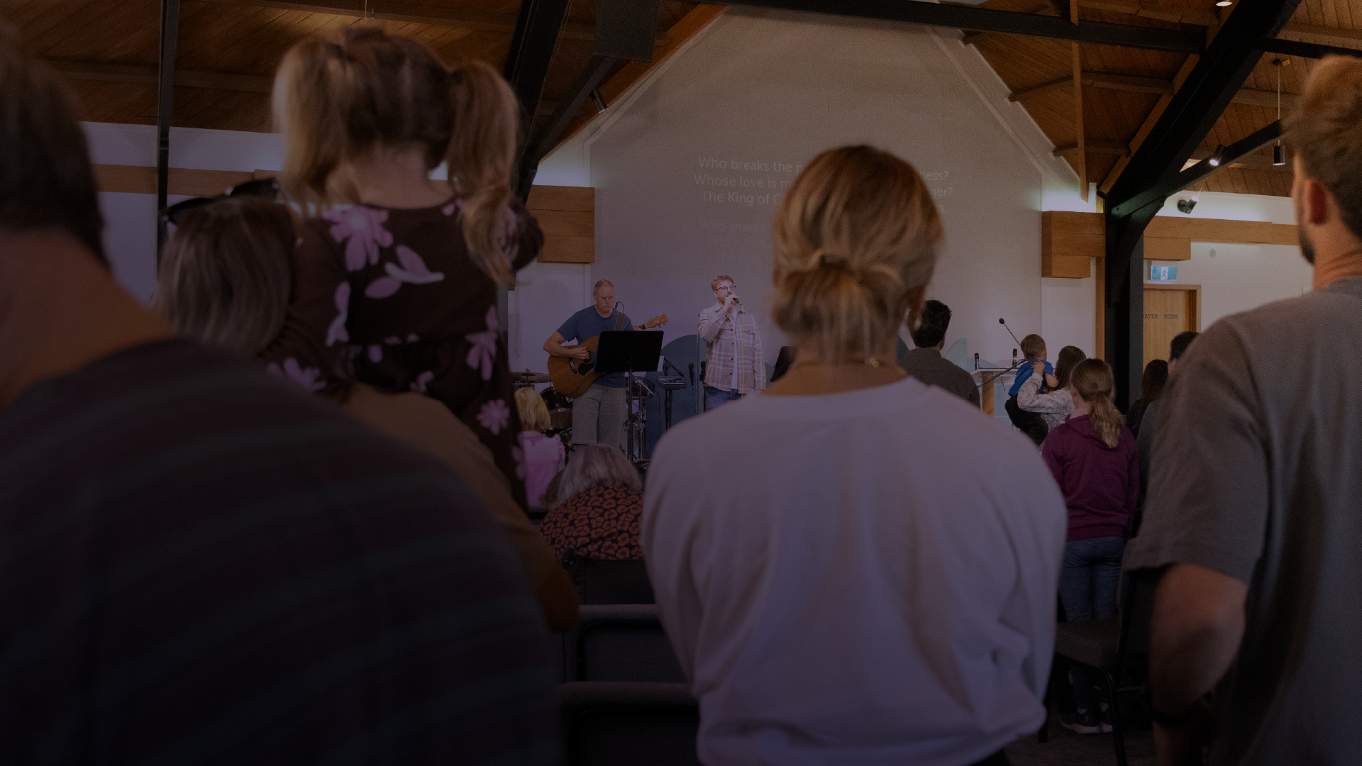 People standing and sitting in a church or auditorium listening to a speaker and a musician on stage.