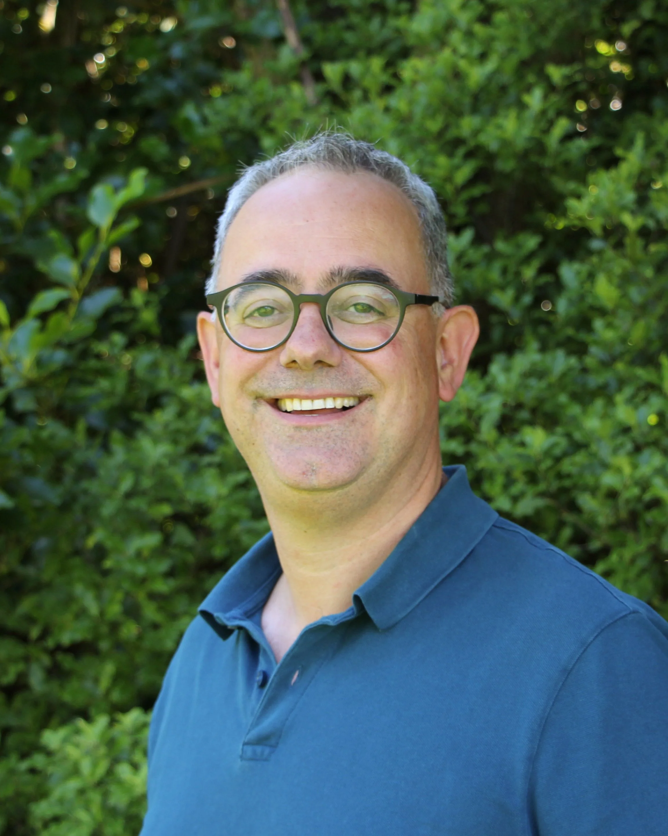Close-up of a smiling man with glasses and gray hair, wearing a blue shirt, outdoors with greenery in the background.