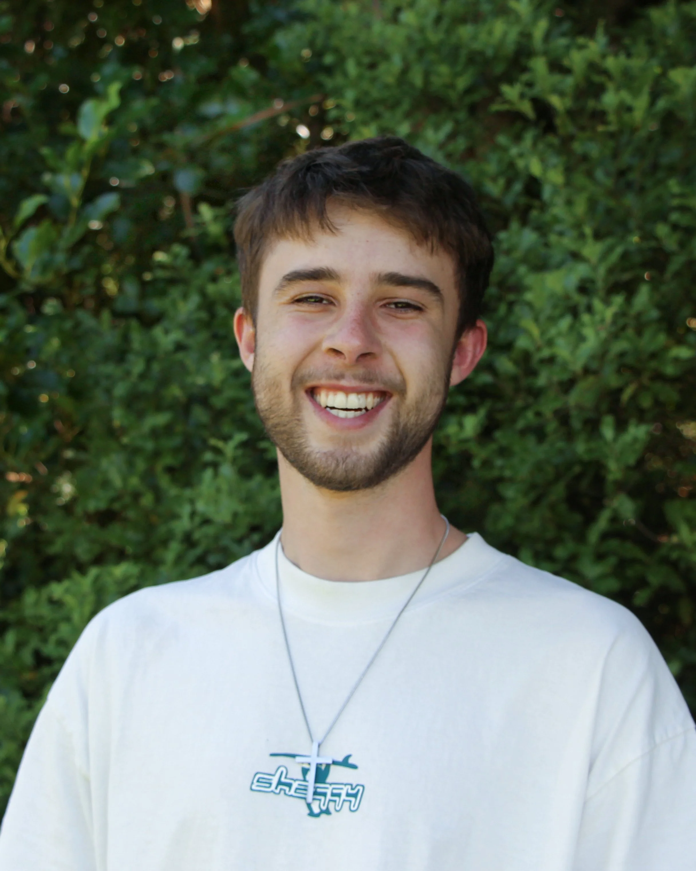 A young man with short dark hair and a beard smiling outdoors against a background of green leaves, wearing a white T-shirt with a graphic design and a necklace with a cross pendant.