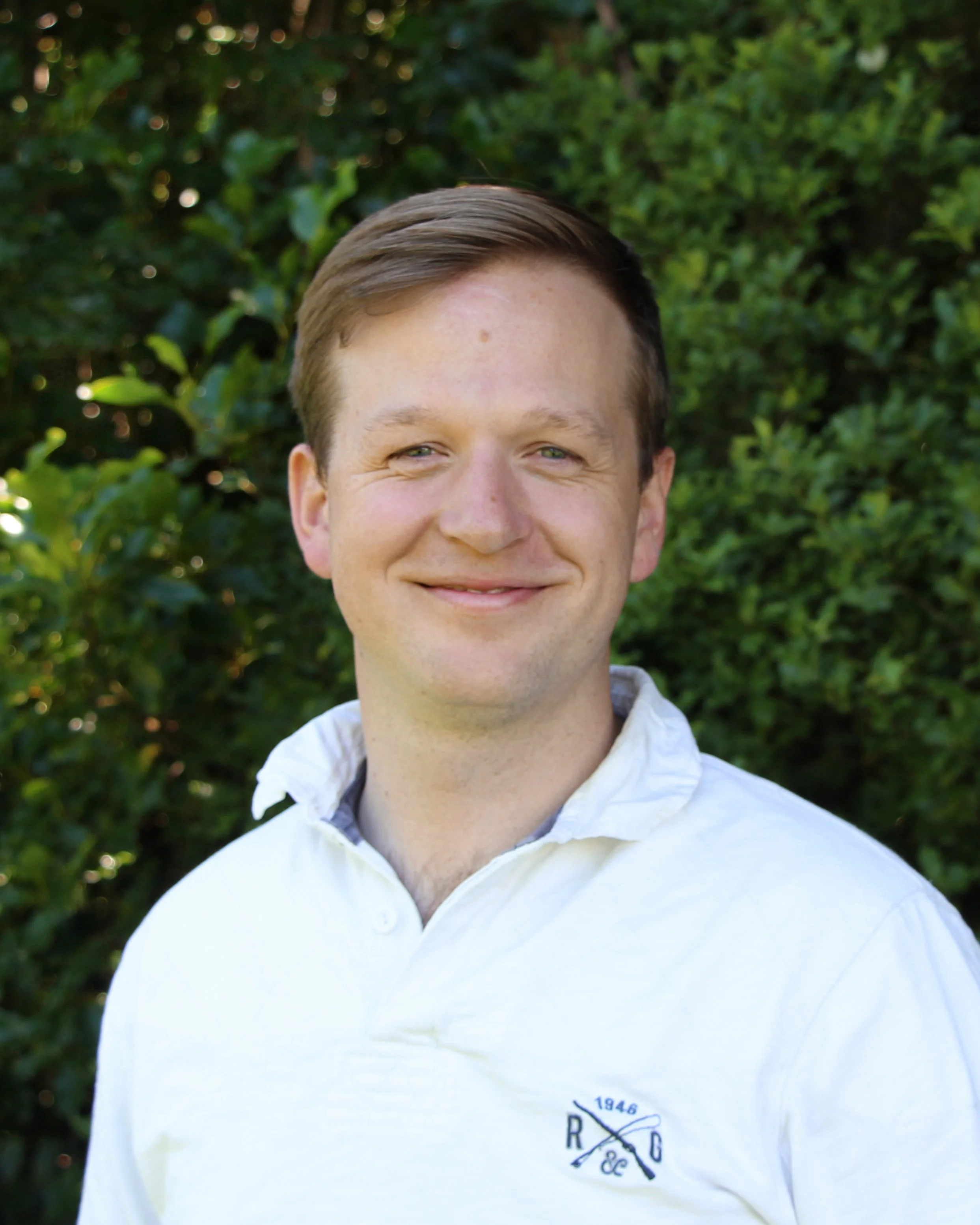 Close-up of a smiling young man with short brown hair, wearing a white collared shirt, standing outdoors in front of green foliage.