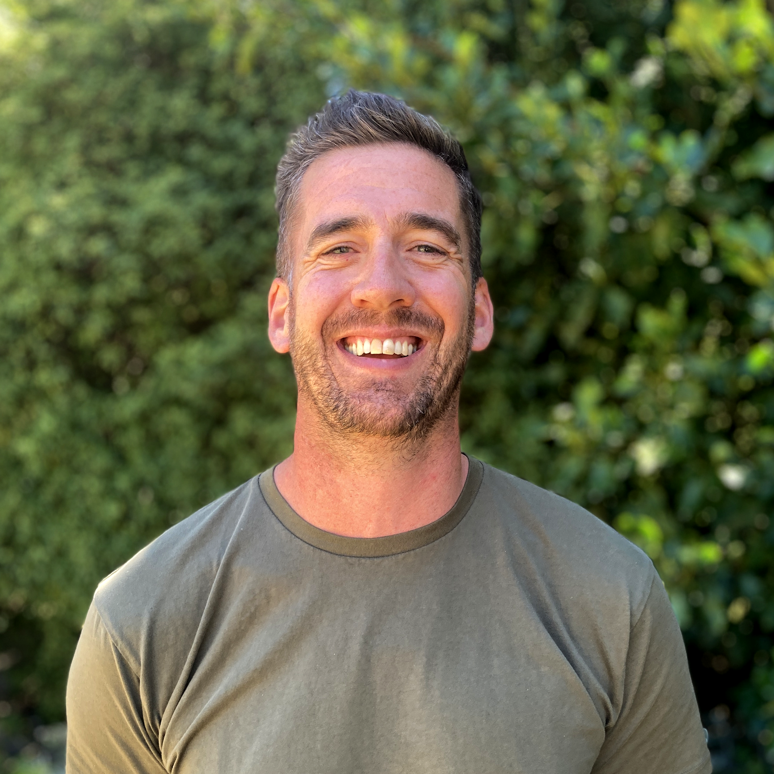 Close-up of a smiling man with a beard and short hair, wearing a gray t-shirt, outdoors with green foliage in the background.