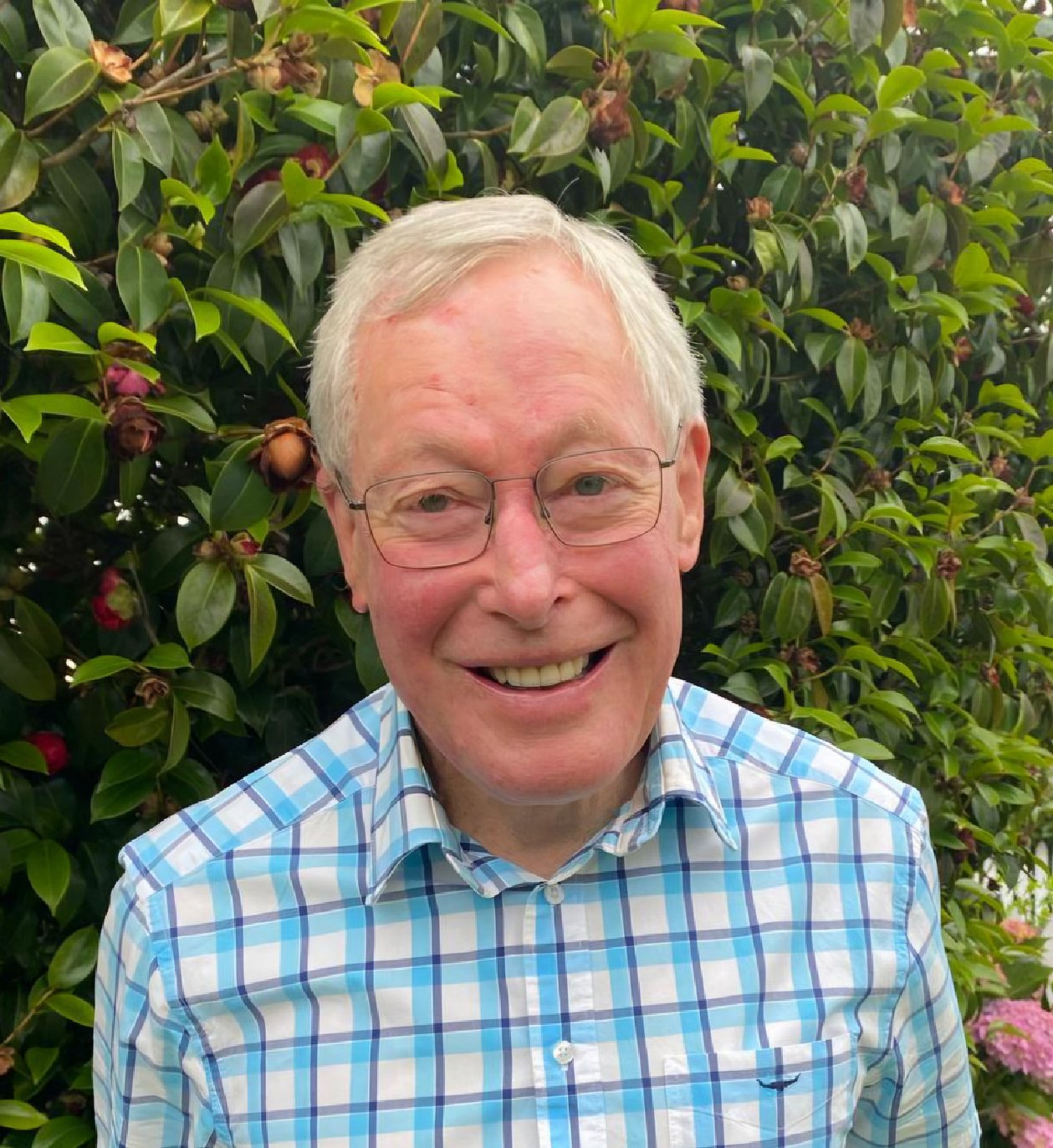 Older man with glasses smiling outdoors in front of green shrub with pink flowers, wearing a blue and white checkered shirt.